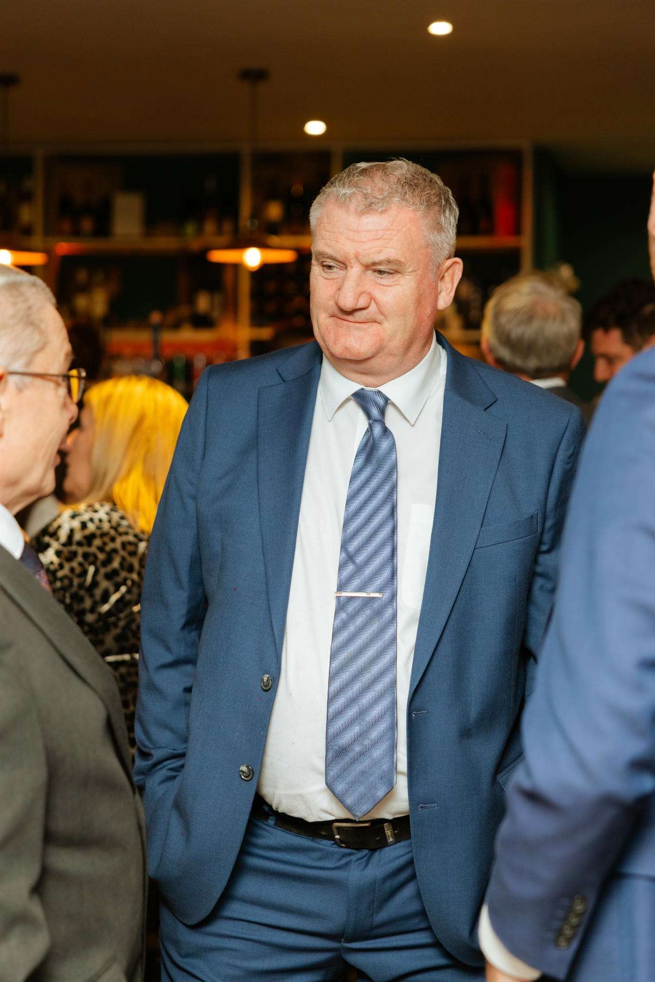 A man in a blue suit with a striped tie stands with his hands in his pockets at a social gathering.