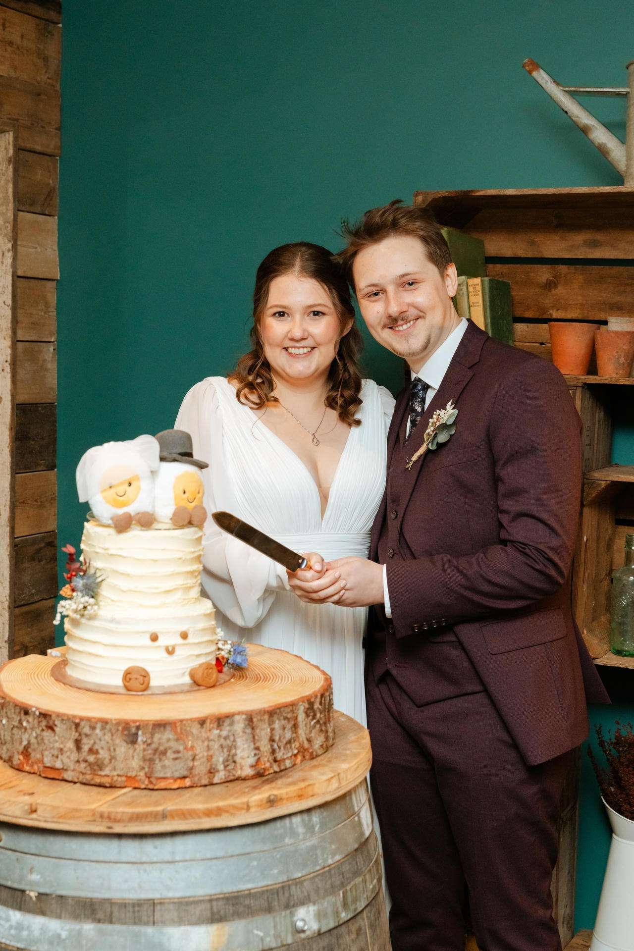 Smiling bride in a white dress and groom in a burgundy suit cut a rustic two-tier wedding cake topped with sheep toppers, standing beside a wooden slab.