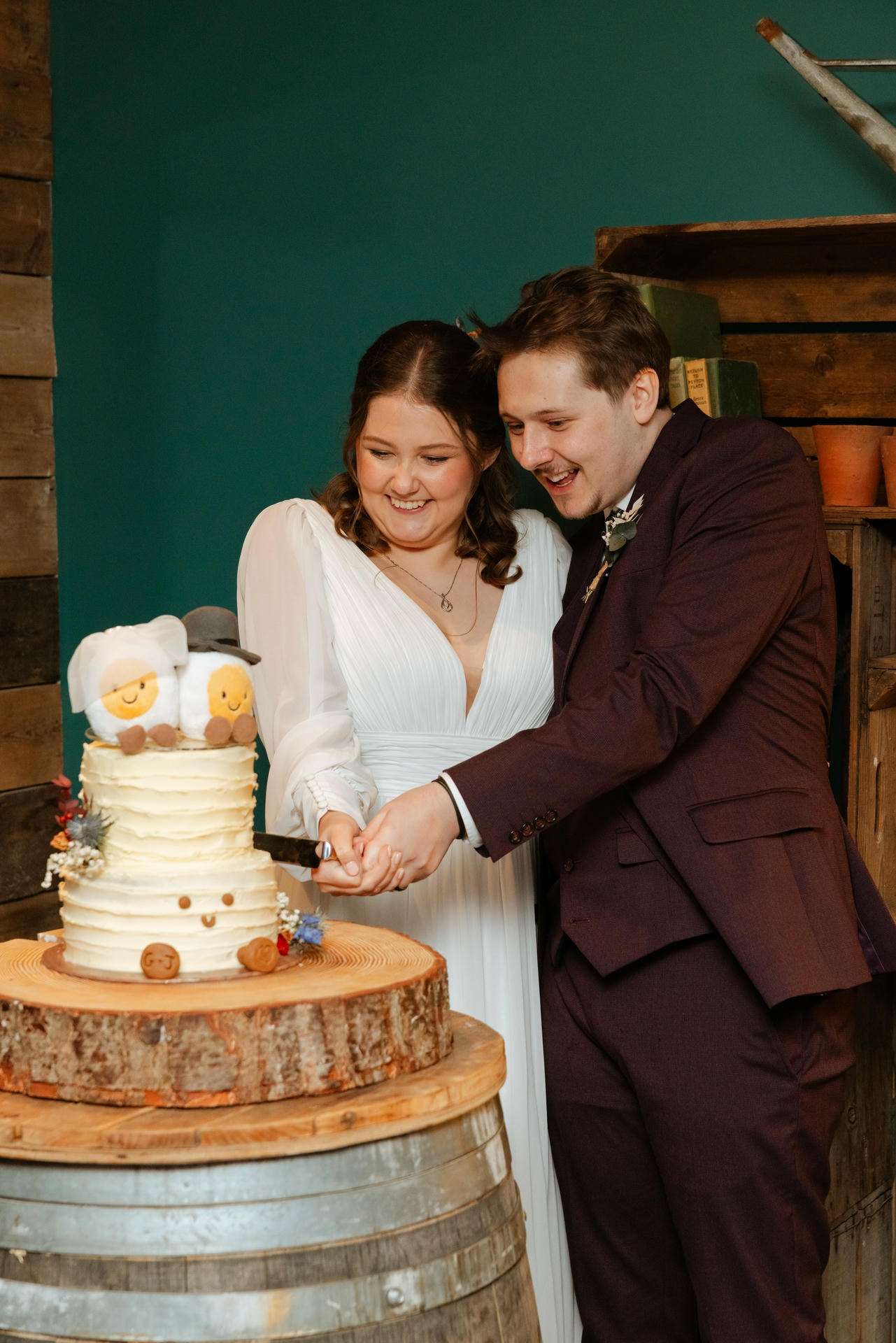 A bride and groom in wedding attire cut a white-frosted wedding cake with plush sheep toppers on a rustic wooden stand.