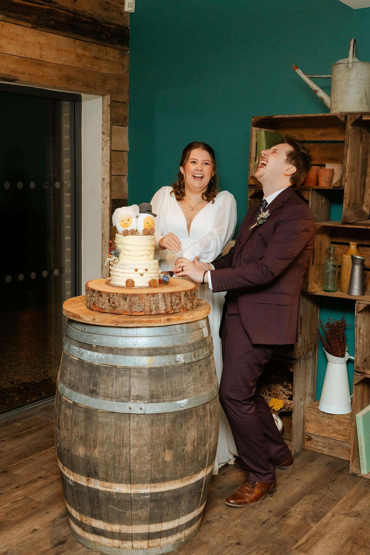 Bride and groom cutting a two-tier wedding cake topped with teddy bears on a wooden barrel in a rustic venue.