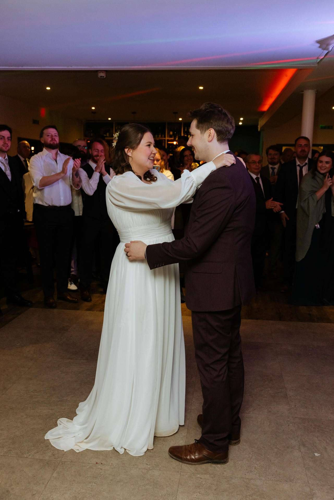 Bride in a white wedding dress dances with a groom in a dark suit as guests applaud at the wedding reception.