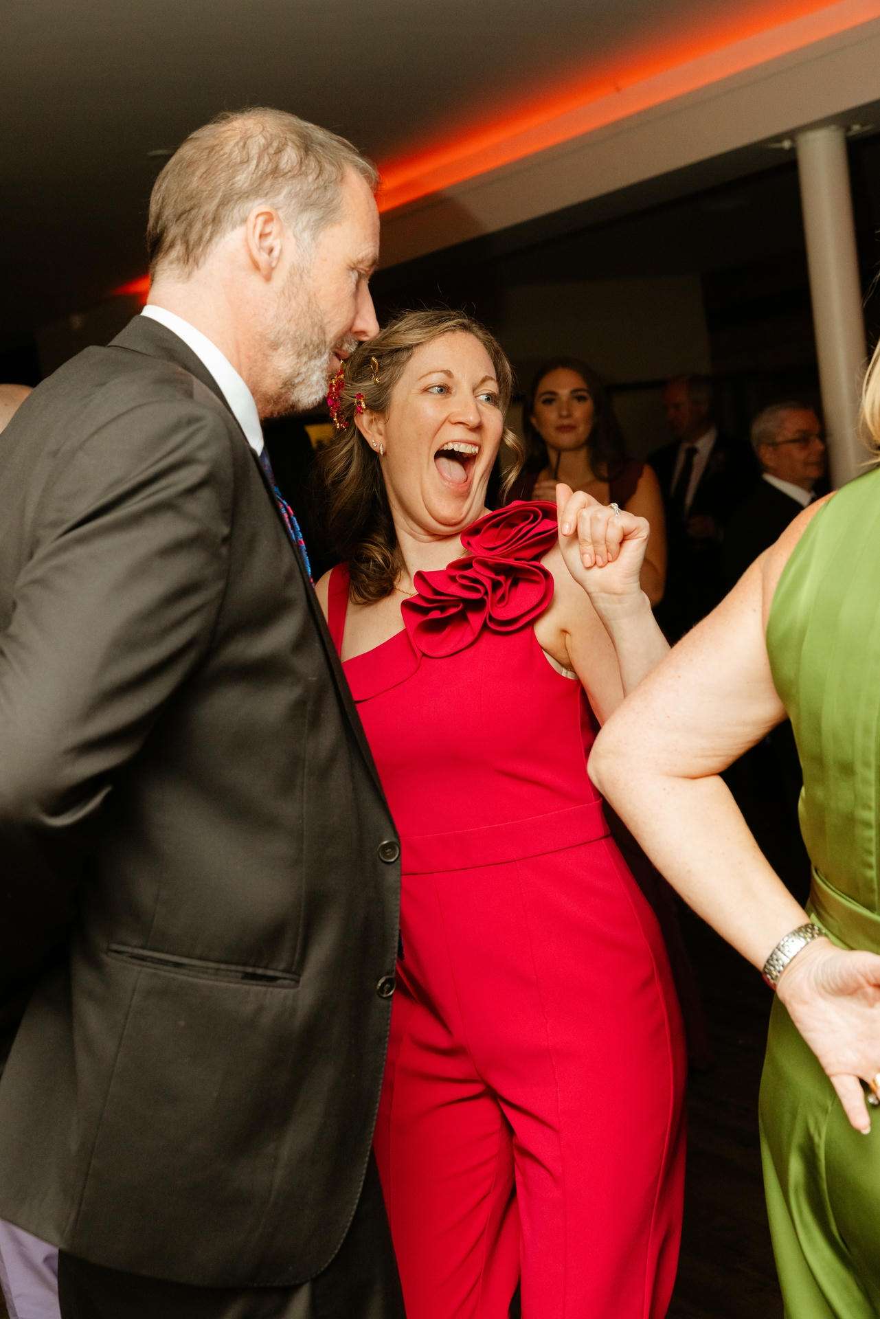 A woman in a bright red dress with a large floral shoulder detail laughs and dances with a gray‑haired man in a dark suit at a party.