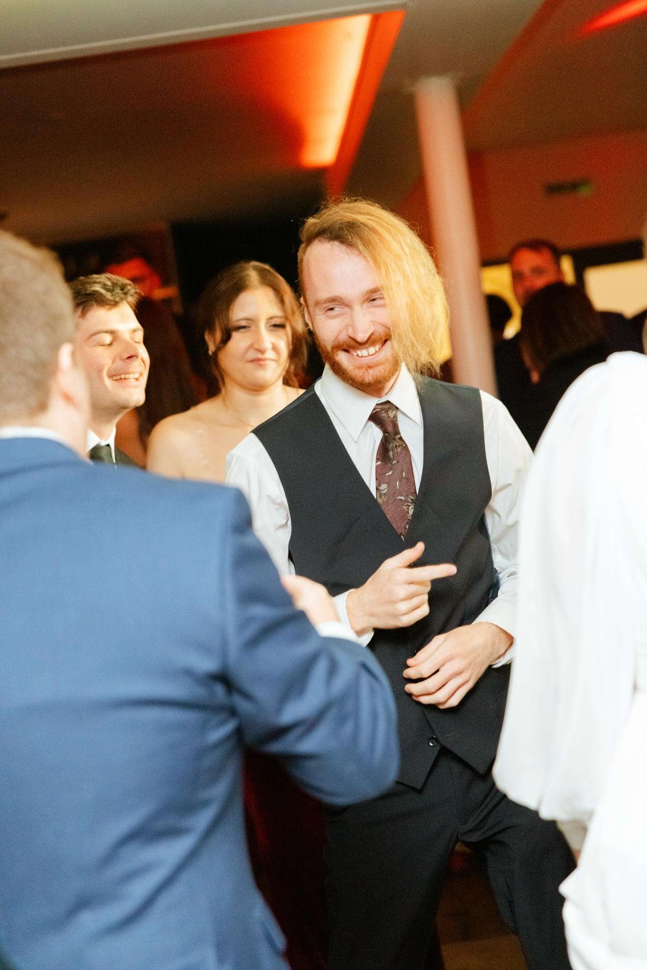 A man with shoulder-length blond hair wearing a white shirt and black vest dances and smiles at a party.