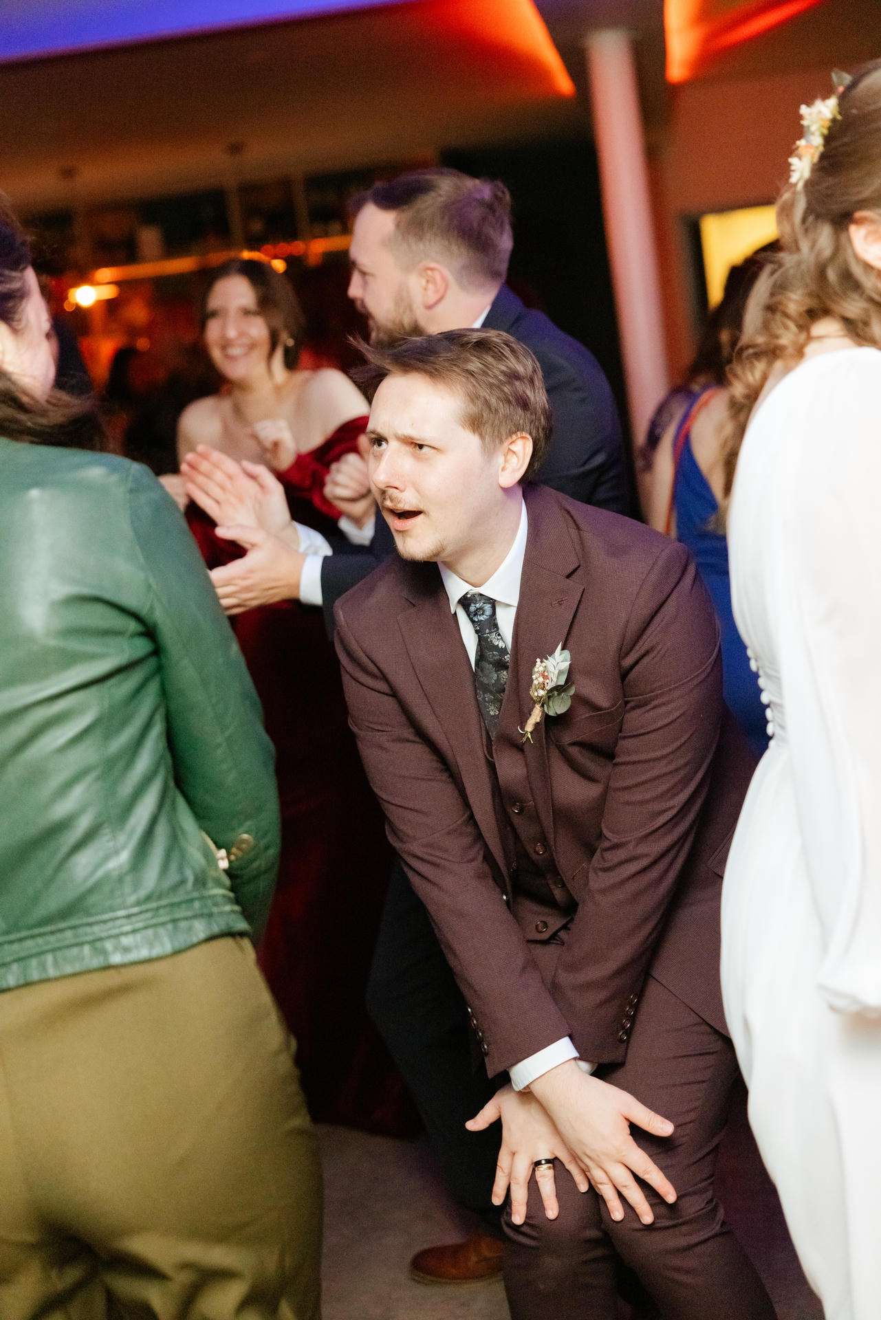 A man in a brown suit dancing at a lively party, surrounded by guests clapping.