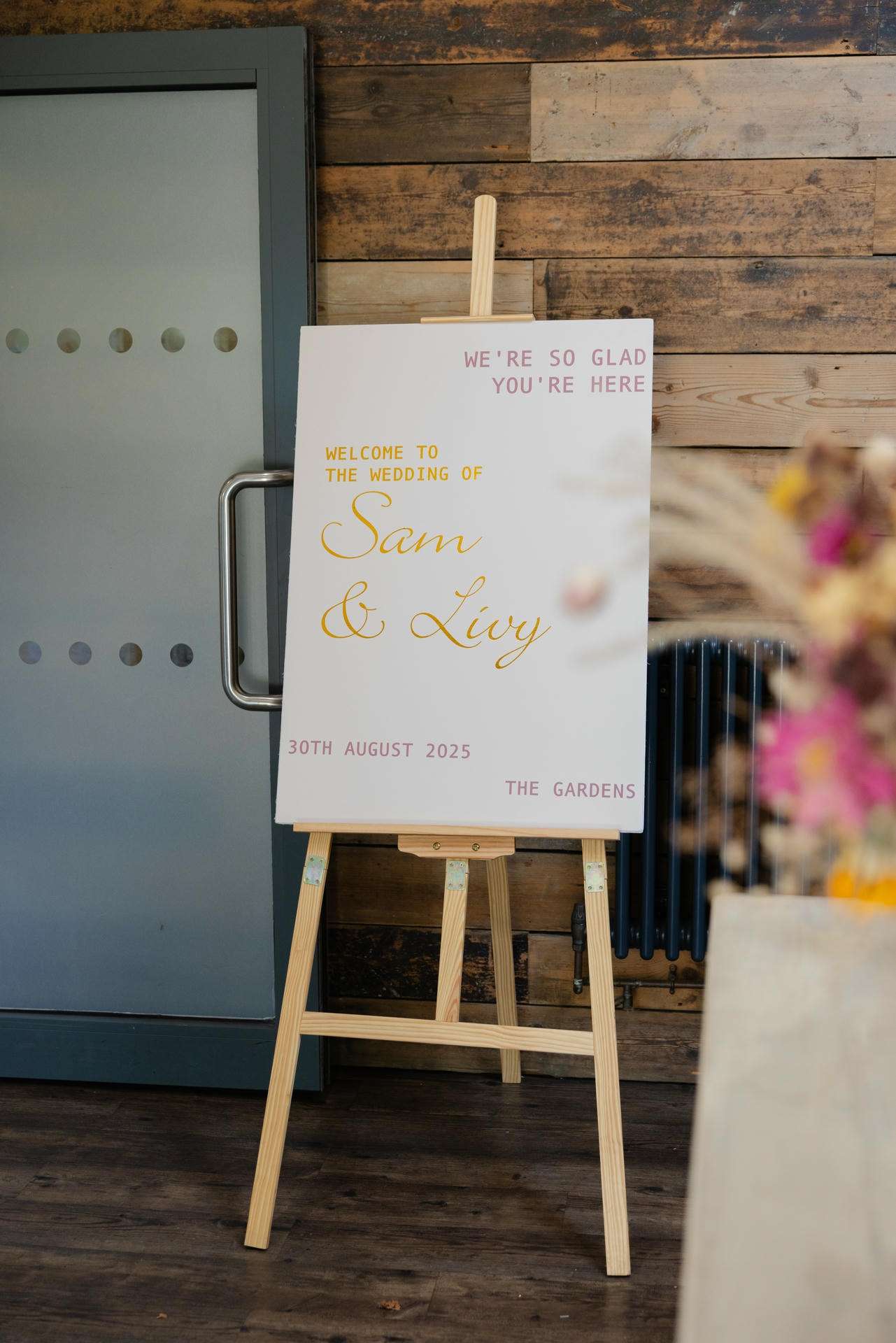A wedding welcome sign on a wooden easel against a rustic wooden wall, with gold calligraphy for Sam & Livy and pink text reading "We're so glad you're here."