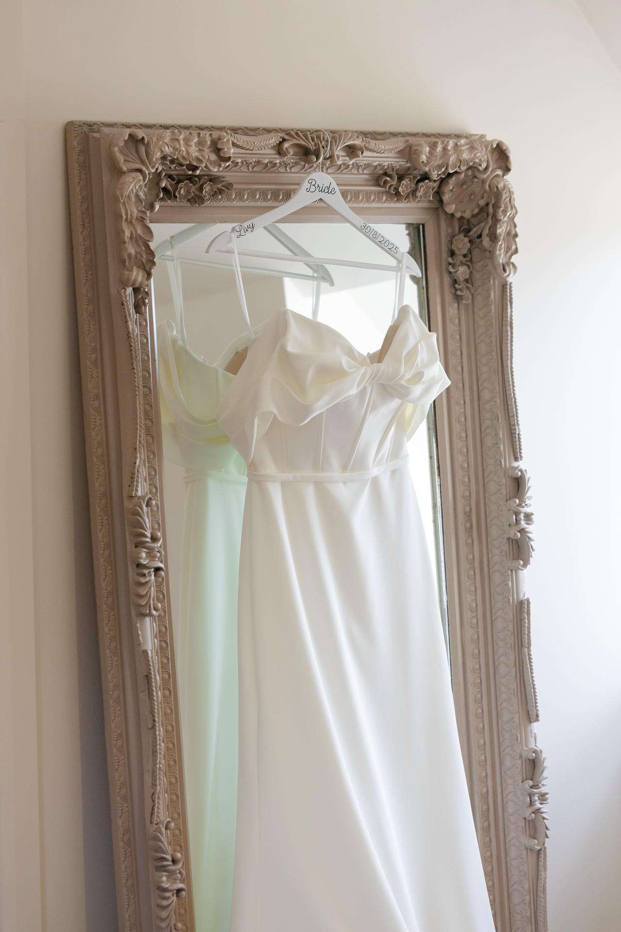 White wedding dress on a hanger labeled Bride hangs in front of an ornate beige-framed mirror.