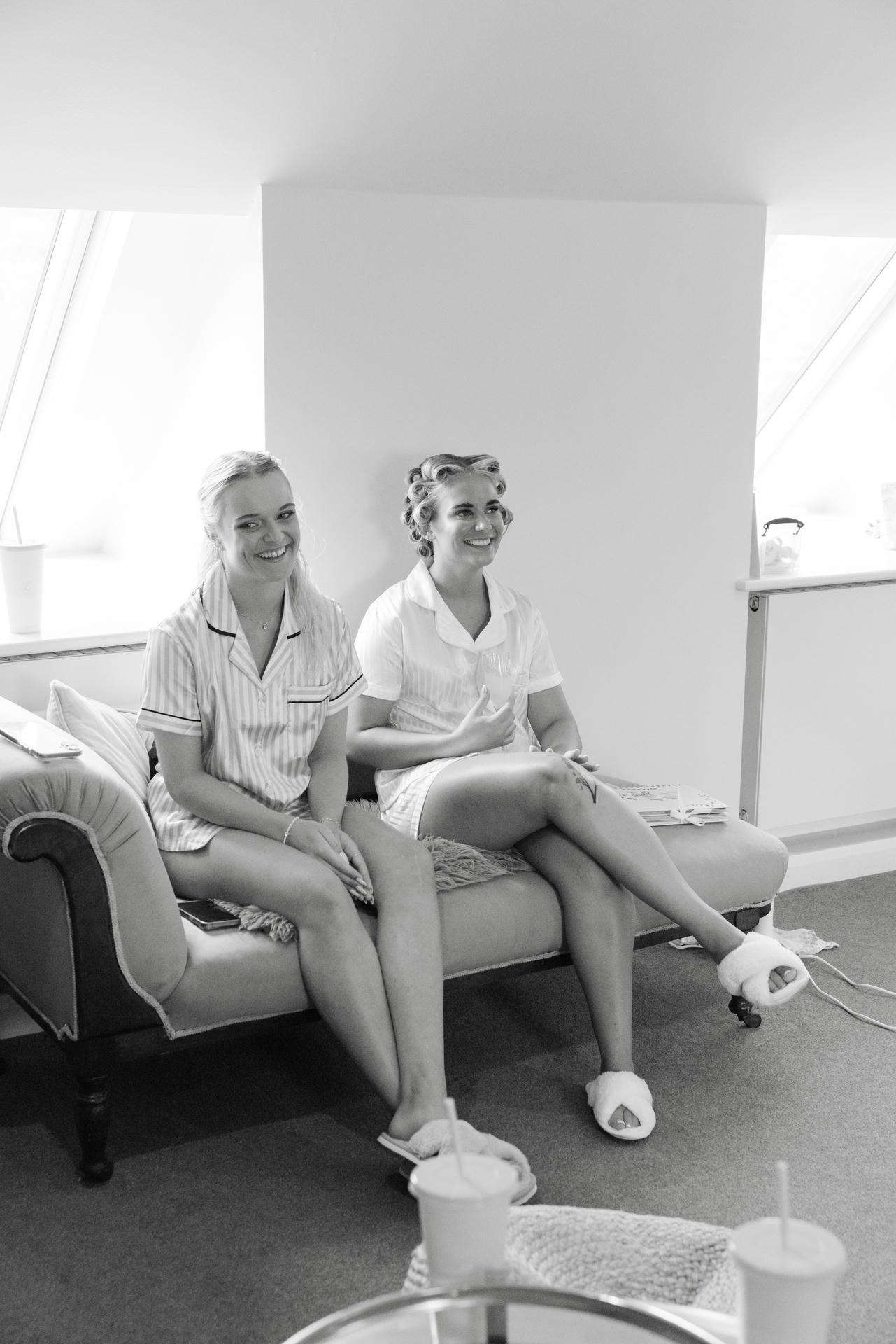 Two women in pajamas with hair curlers sit on a sofa in a bright room, smiling.