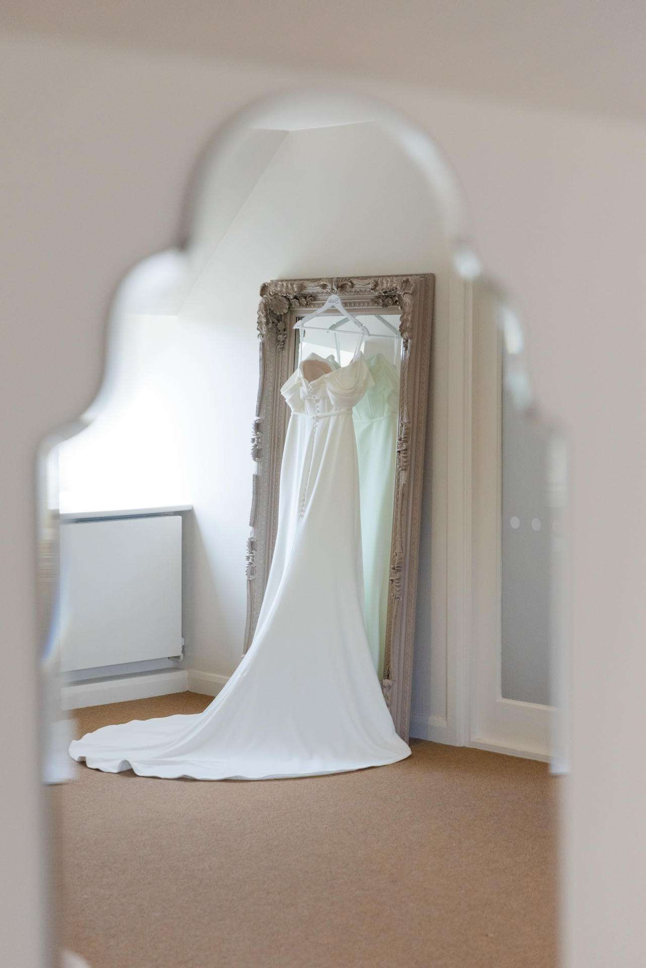 A white wedding dress on a hanger is reflected in an ornate vintage mirror, with a long train flowing onto the beige carpet.