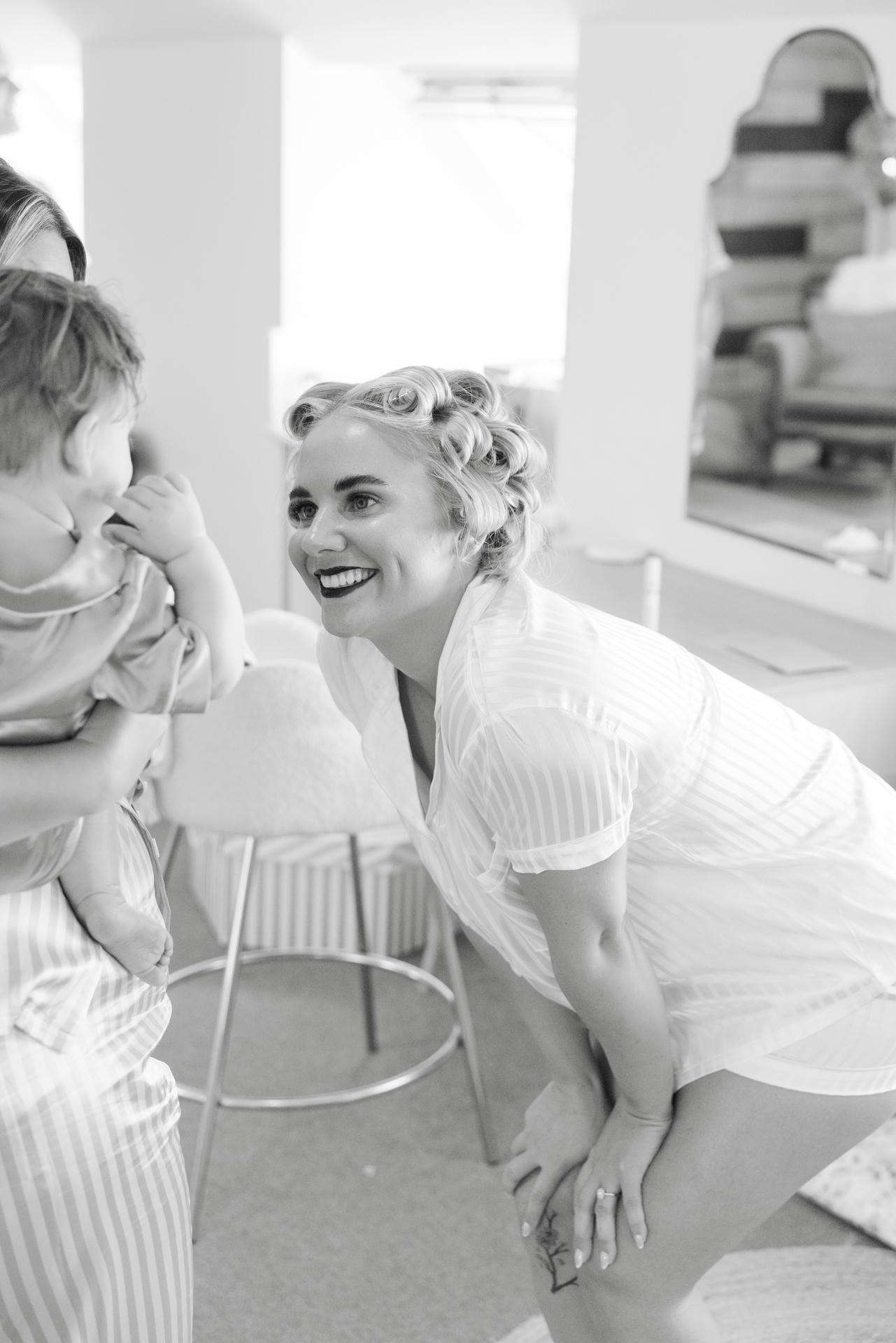 A smiling woman with curlers in her hair bends toward a baby being held by another person in a bright room.
