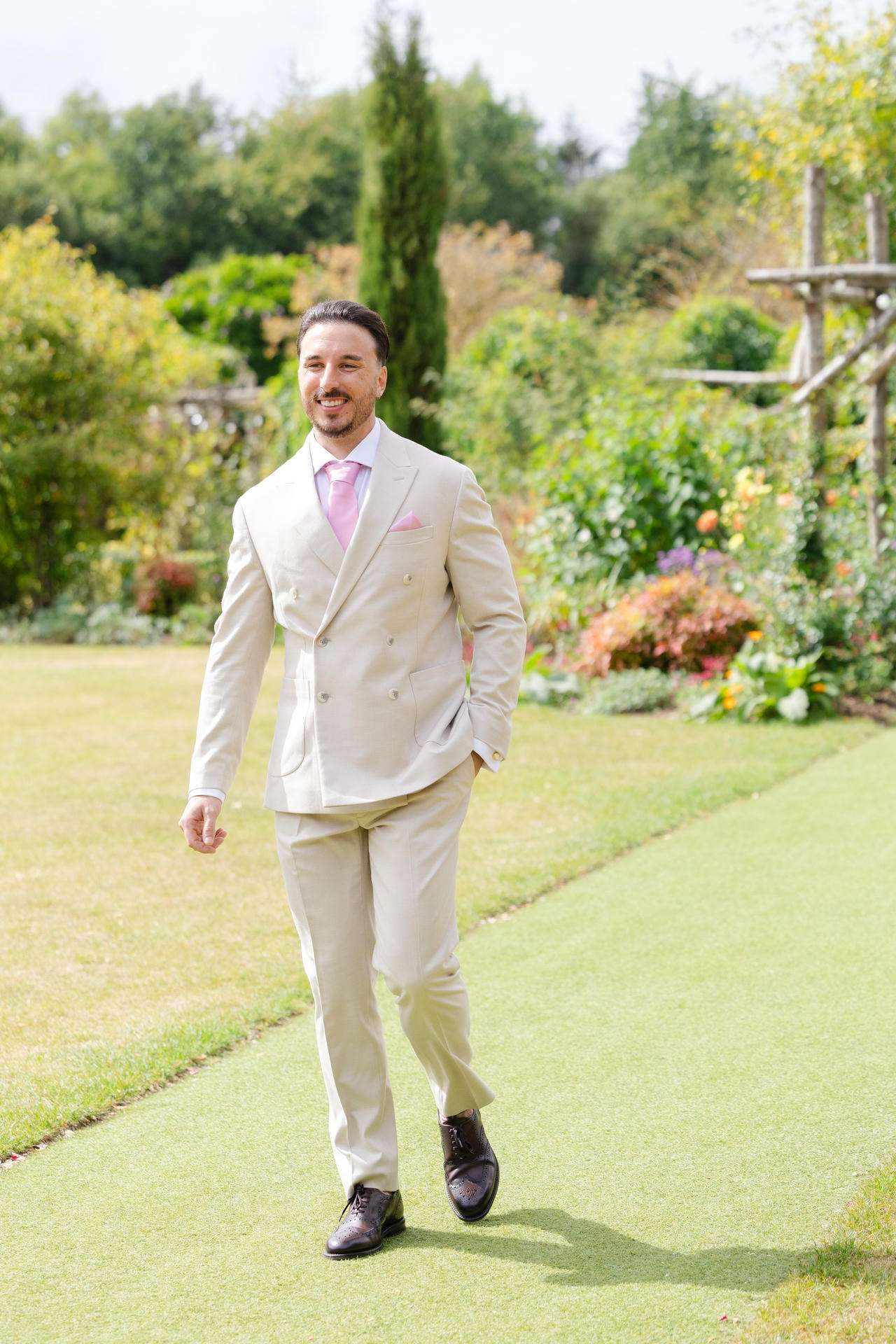 A man in a light beige suit with a pink tie and pocket square walks along a garden path surrounded by greenery.