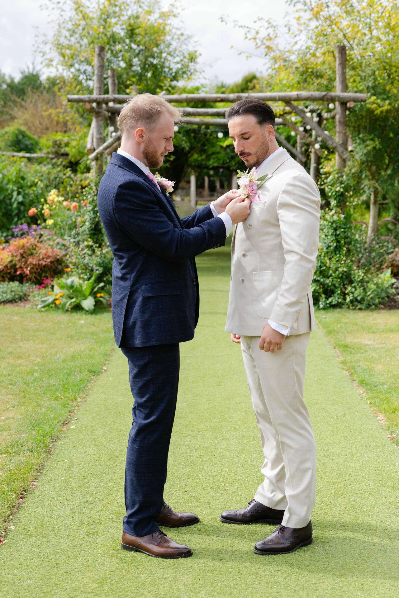 Two men in suits stand on a garden path while one pins a pink boutonniere onto the other's cream-colored suit.