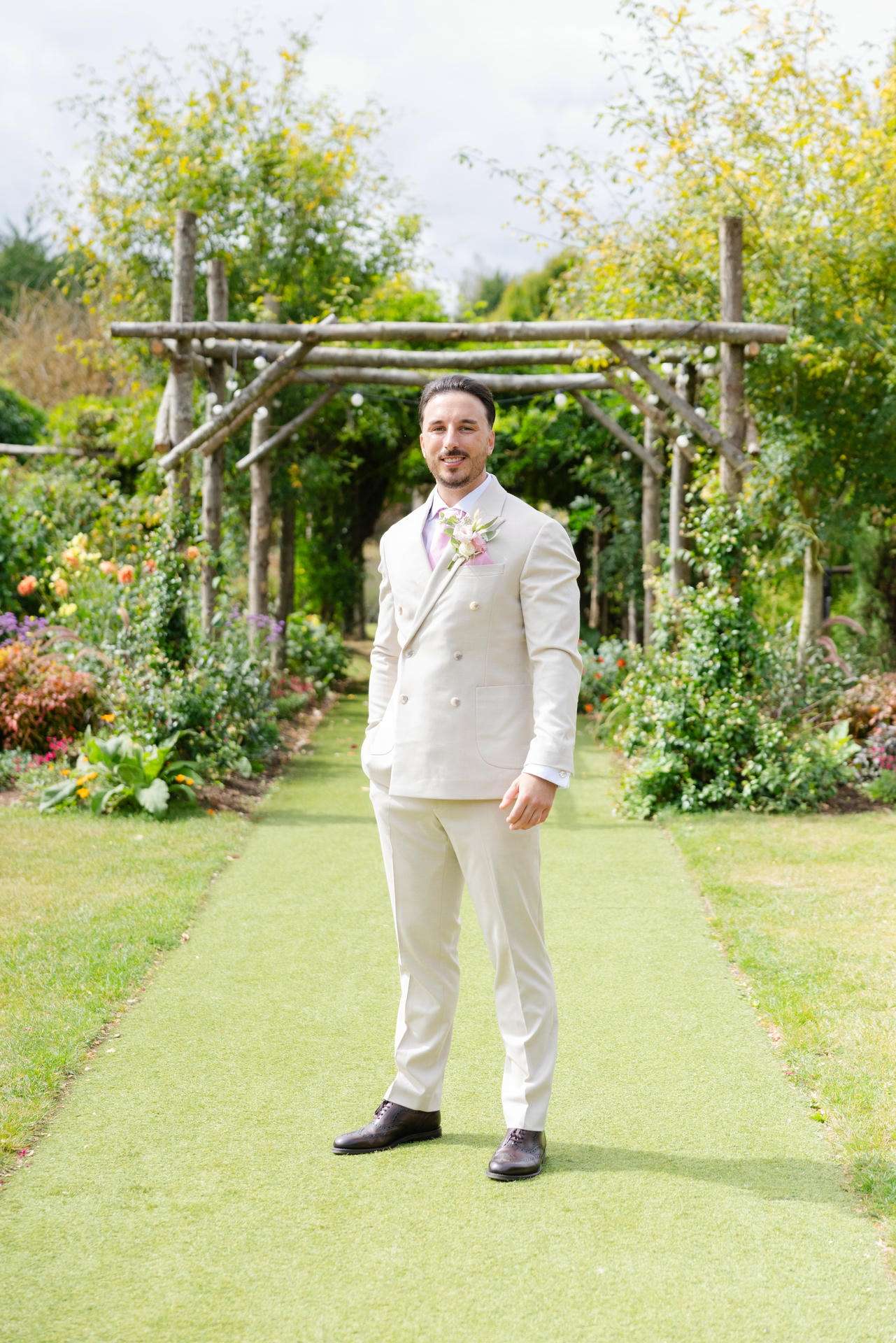 A man in a light beige suit stands on a garden path under a wooden arbor, surrounded by colorful flowers.