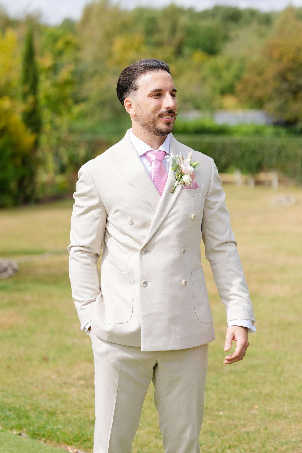 A man wearing a light beige double-breasted suit with a pink tie and floral boutonniere stands outdoors in a grassy garden.