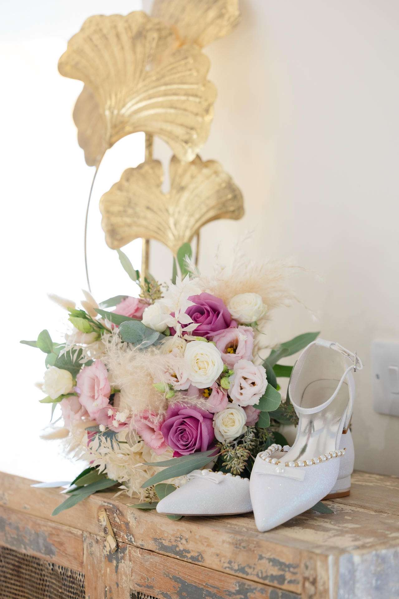 A pastel bridal bouquet of pink and white roses with greenery rests beside white satin wedding shoes adorned with pearls on a weathered wooden surface.
