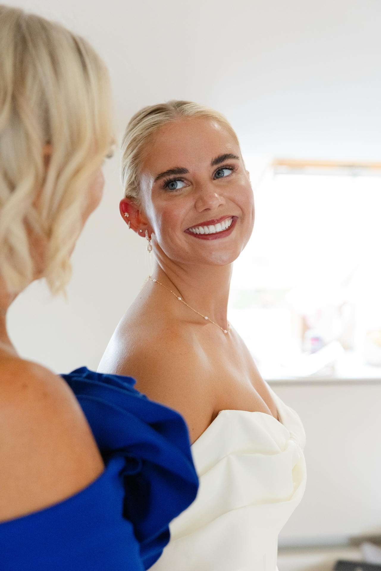 Smiling bride with blonde hair in a white strapless wedding dress looks at a woman in a blue dress.