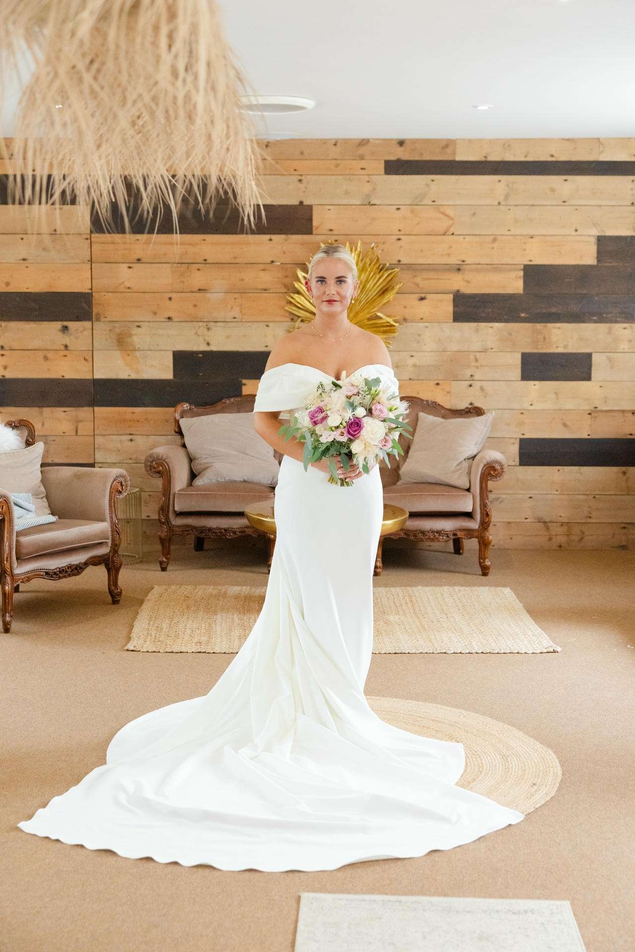 Bride in an off-the-shoulder white wedding gown with a long train, holding a bouquet of pink and white flowers, standing in a rustic wood-paneled room with vintage couches.
