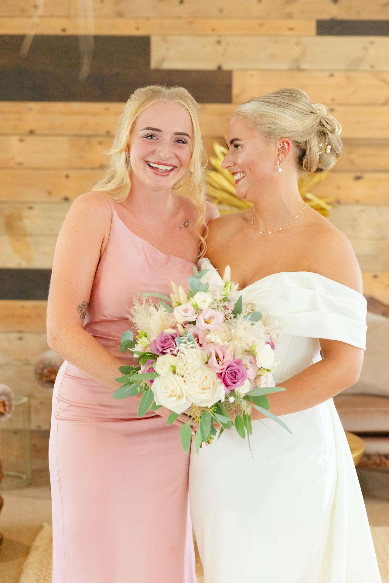 Two women at a wedding, a bride in a white off-shoulder gown holding a pastel bouquet and a bridesmaid in a pink dress smiling together.