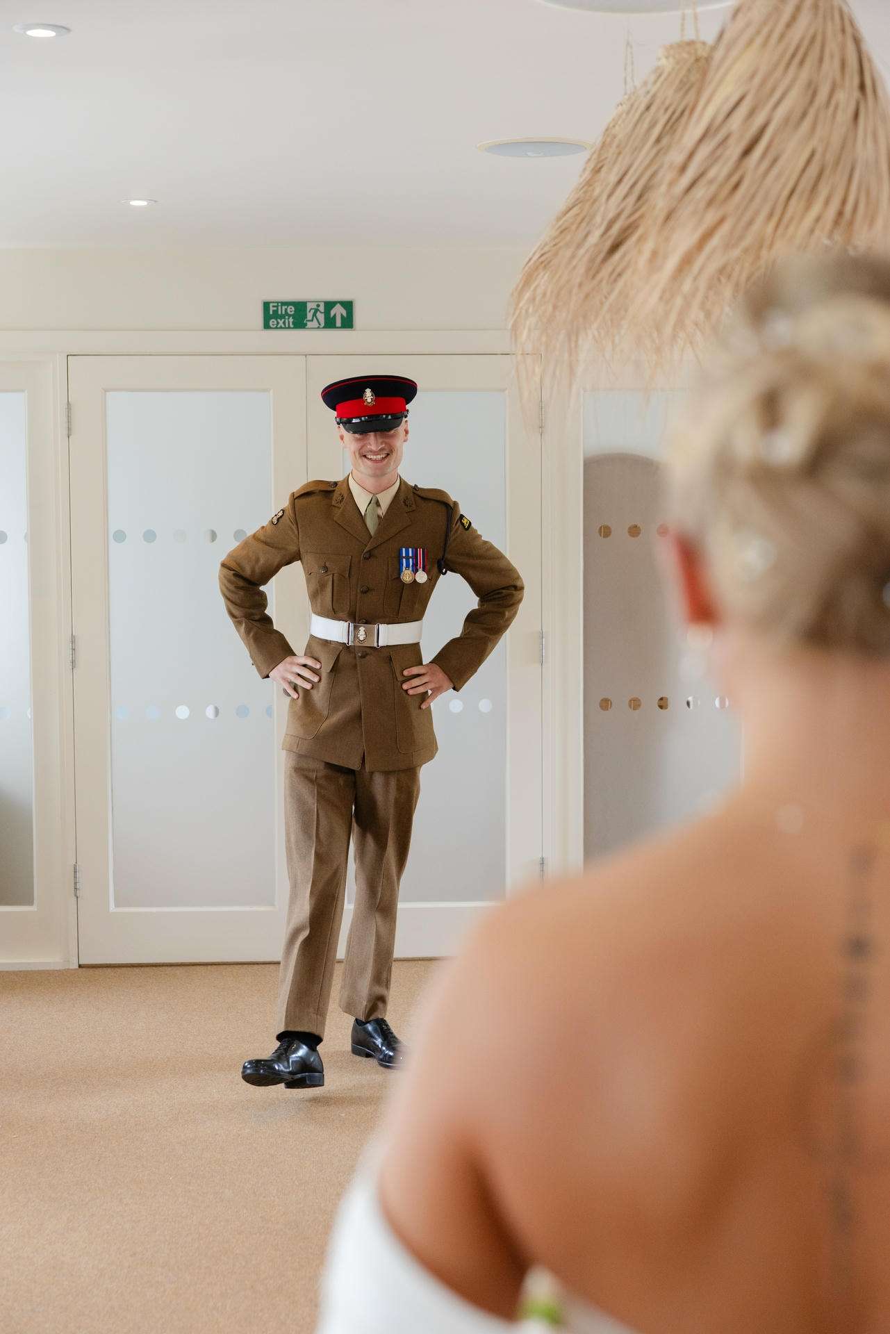 A man in a brown military uniform with medals and a red peaked cap stands with his hands on his hips in a doorway, while a blonde person in the foreground looks on.
