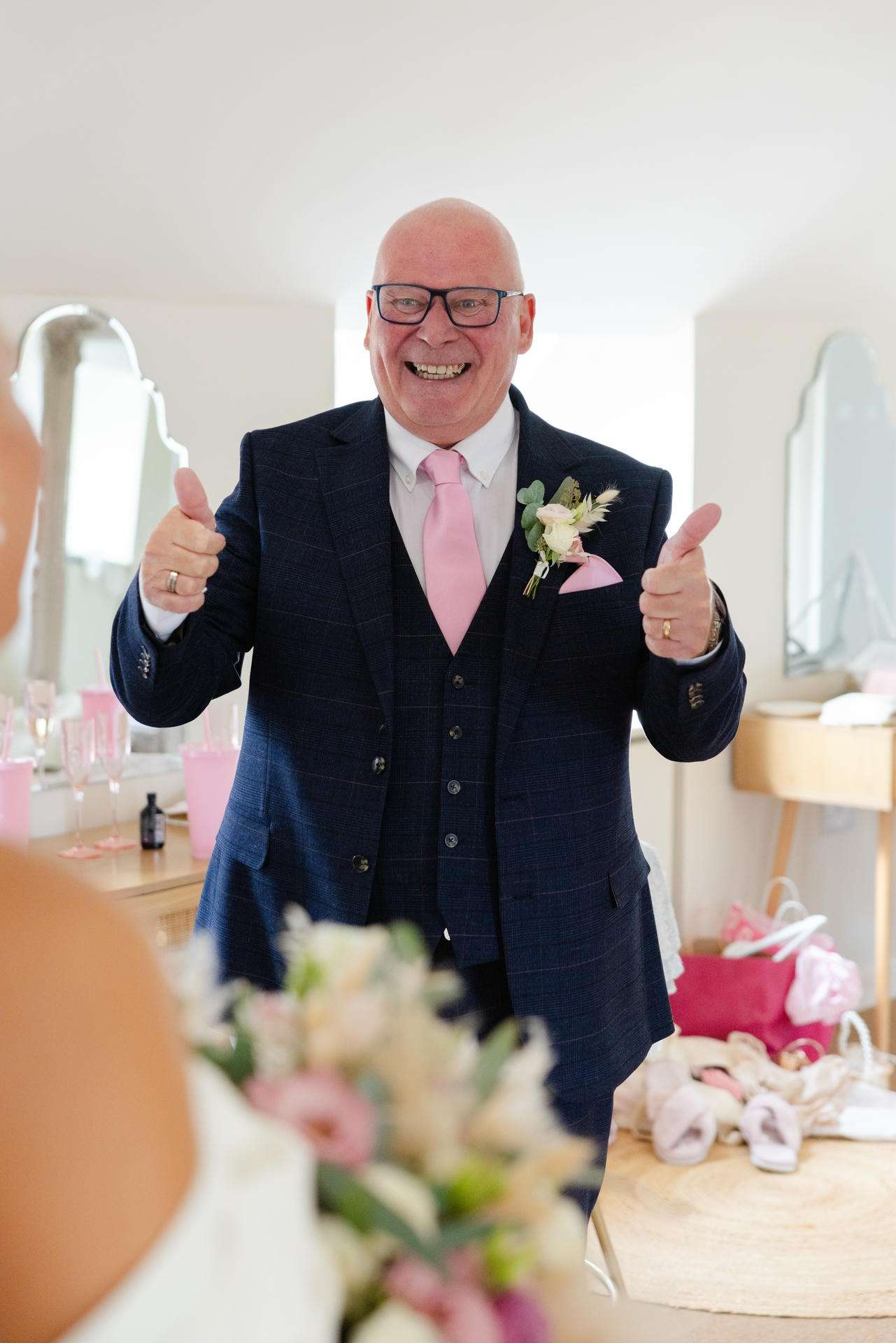 Smiling bald man in a navy suit with a pink tie and boutonniere giving two thumbs up.