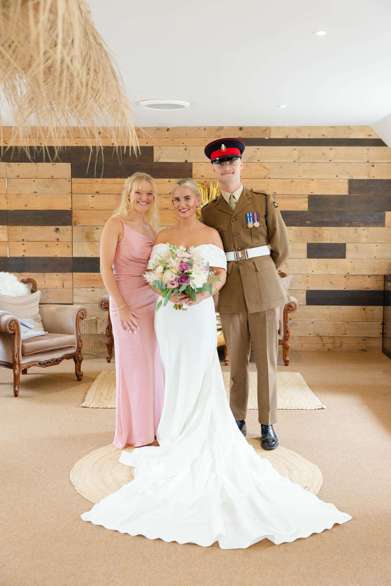 Bride in an off-the-shoulder white wedding gown holding a bouquet stands with a bridesmaid in a pink dress and a soldier in a brown military uniform.