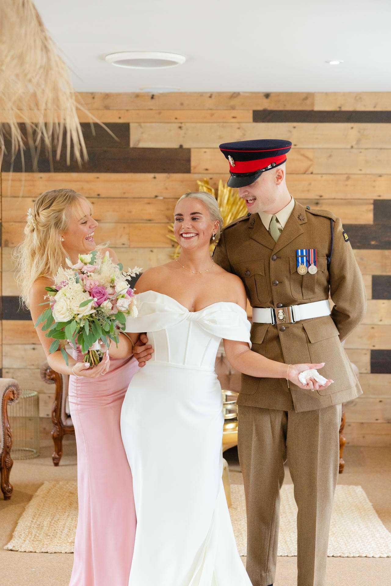 Bride in a white off-the-shoulder wedding gown stands with a smiling bridesmaid in a pink dress and a military officer in dress uniform indoors.