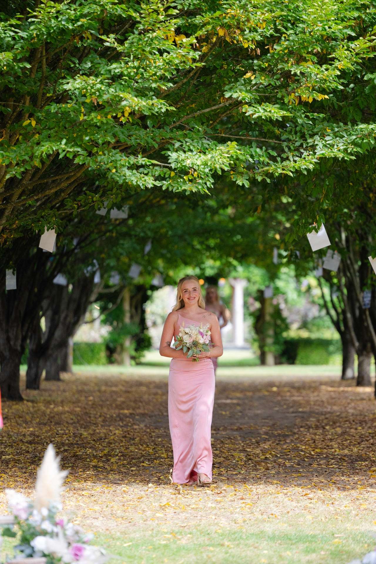 A bridesmaid in a pale pink dress walks down a tree-lined garden path, holding a bouquet.