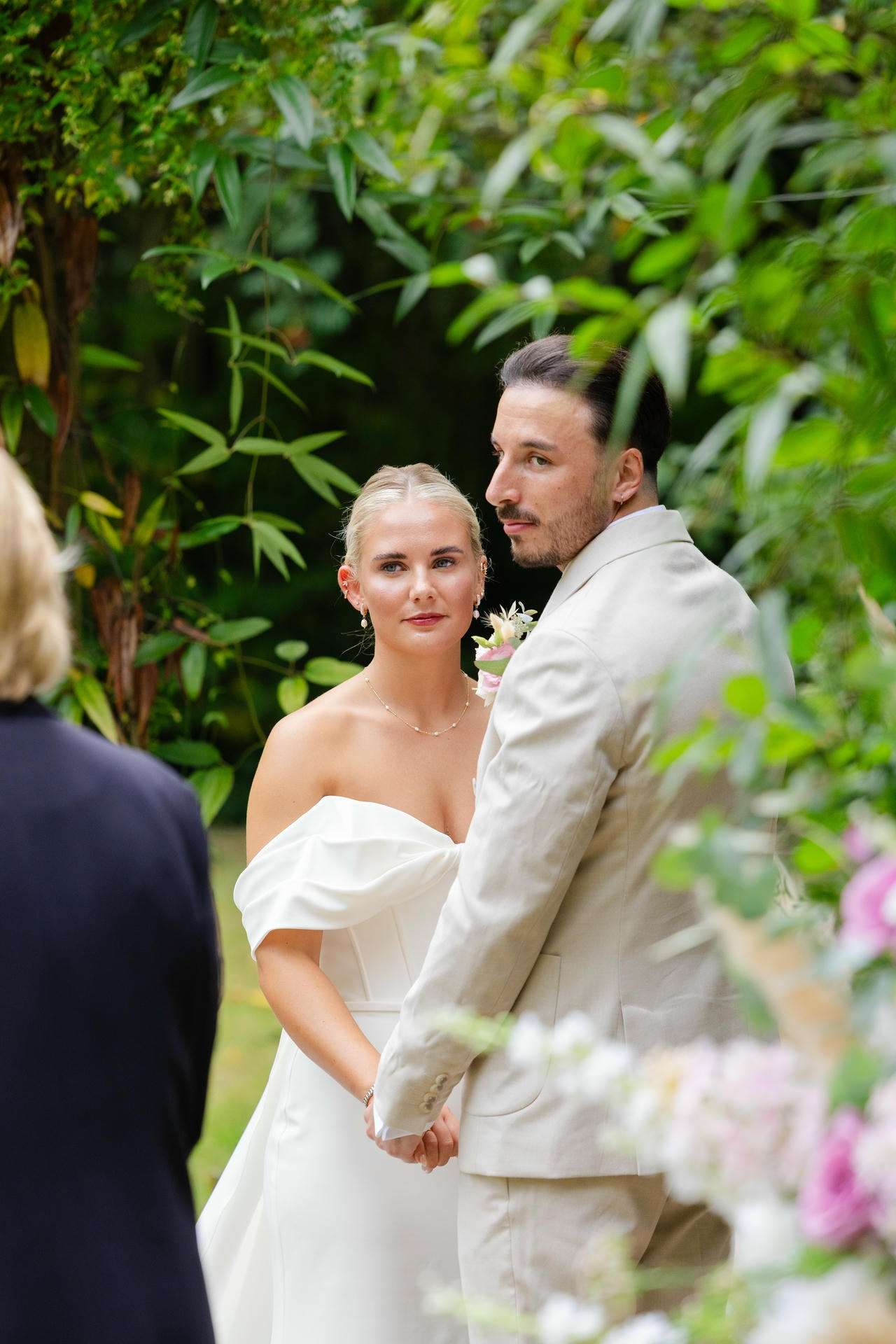 A bride in an off-shoulder white gown and a groom in a light suit hold hands at an outdoor garden wedding.