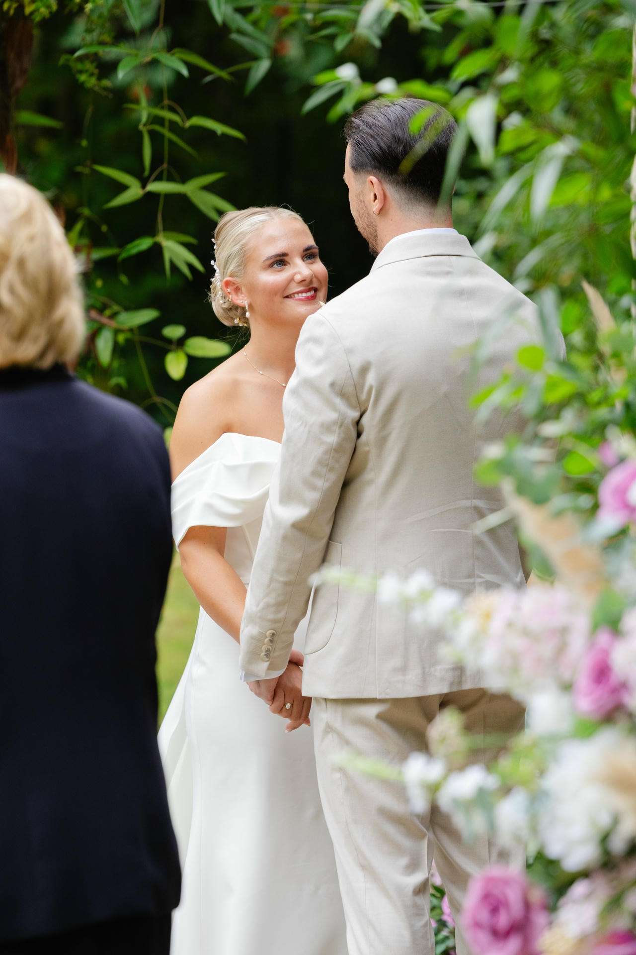 Bride and groom holding hands and gazing at each other during an outdoor wedding surrounded by lush greenery.