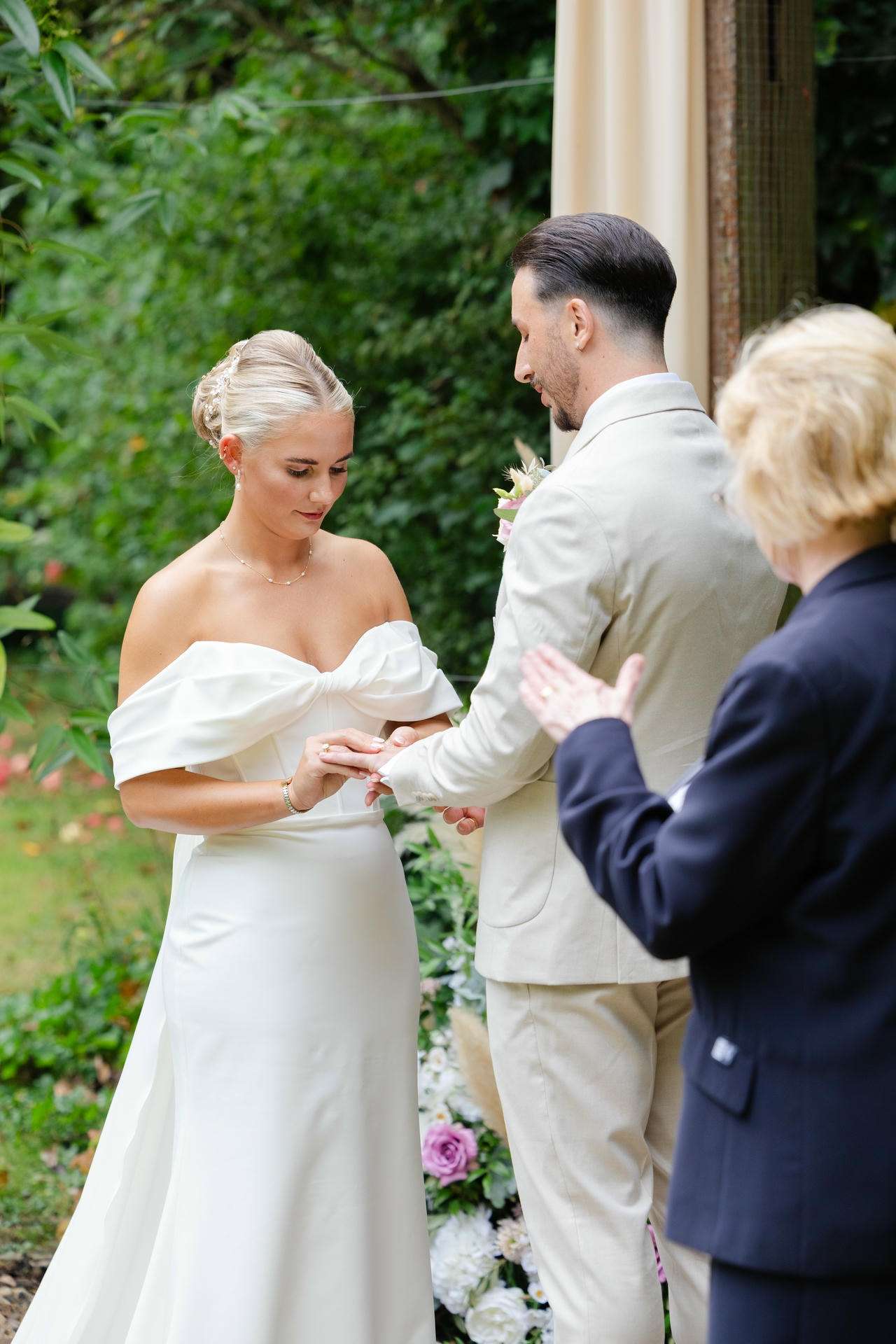 A bride in a white off-shoulder gown places a ring on the groom's finger during an outdoor wedding ceremony.