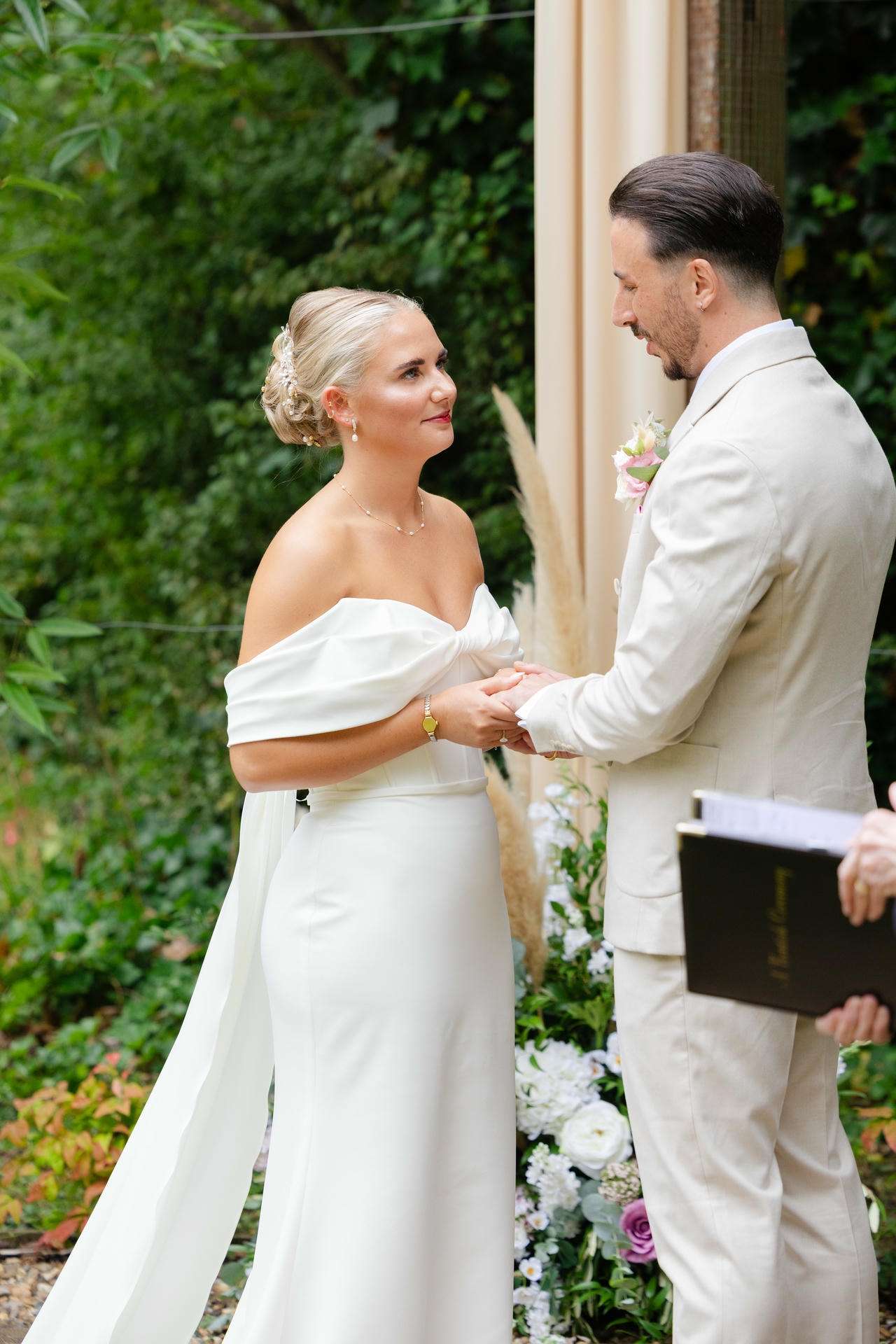 Bride in an off-the-shoulder white gown and a groom in a light suit hold hands during an outdoor wedding ceremony surrounded by greenery and flowers.