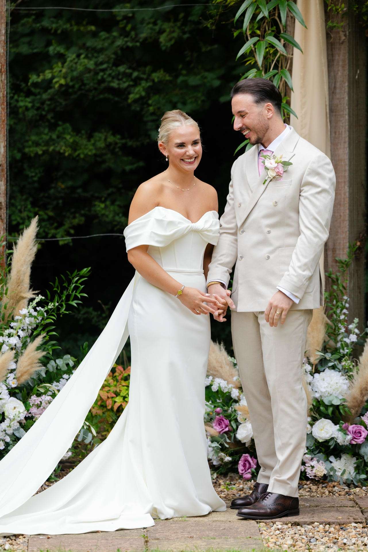 Bride in a strapless white gown with a bow stands with a groom in a light beige suit at an outdoor garden wedding.