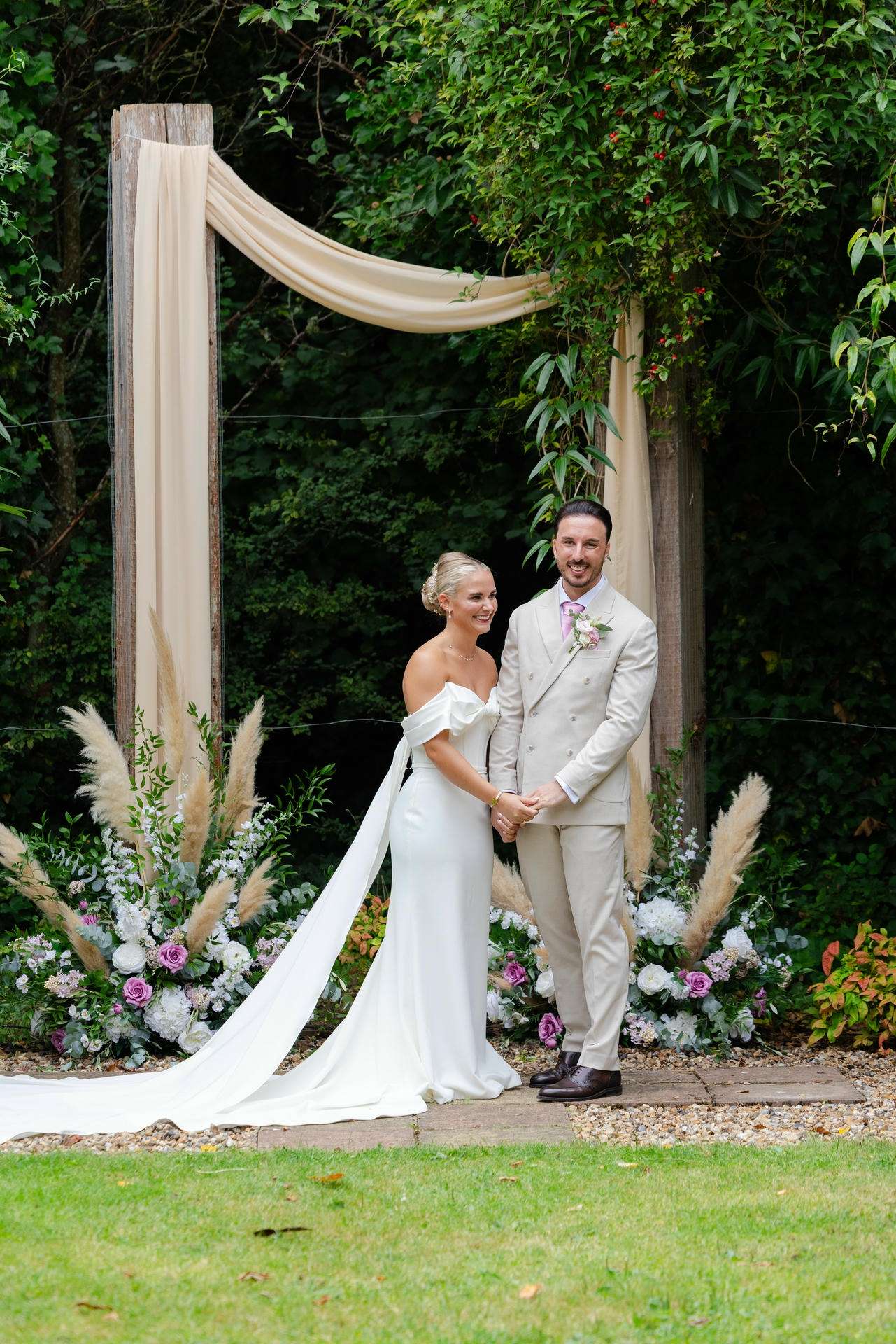 Bride and groom hold hands under a cream fabric arch in a lush garden with pastel floral arrangements.