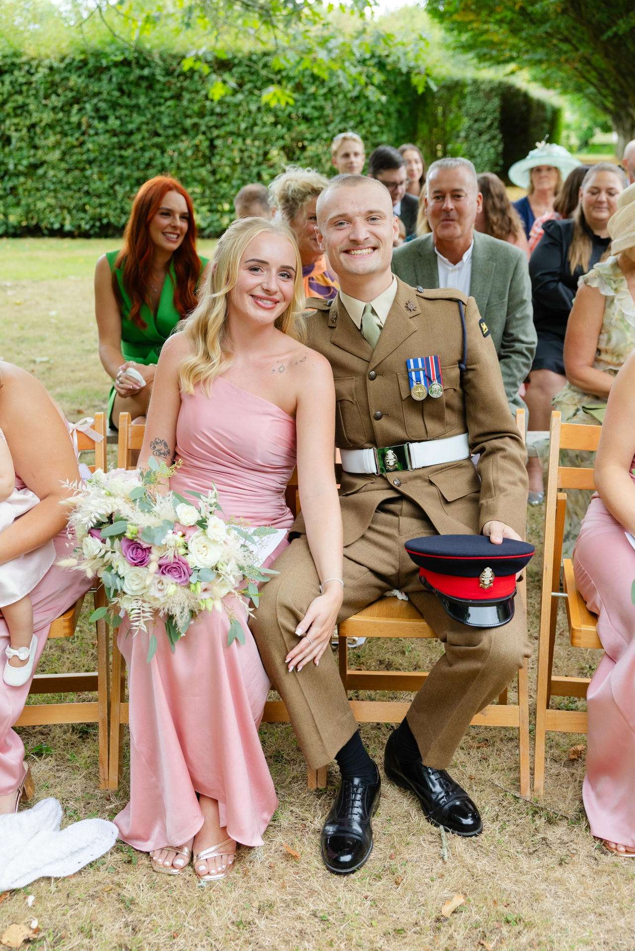 A smiling couple sits on wooden chairs at an outdoor wedding; the woman wears a pink strapless dress and holds a bouquet, while the man in a brown military uniform with medals rests his hat on his lap.