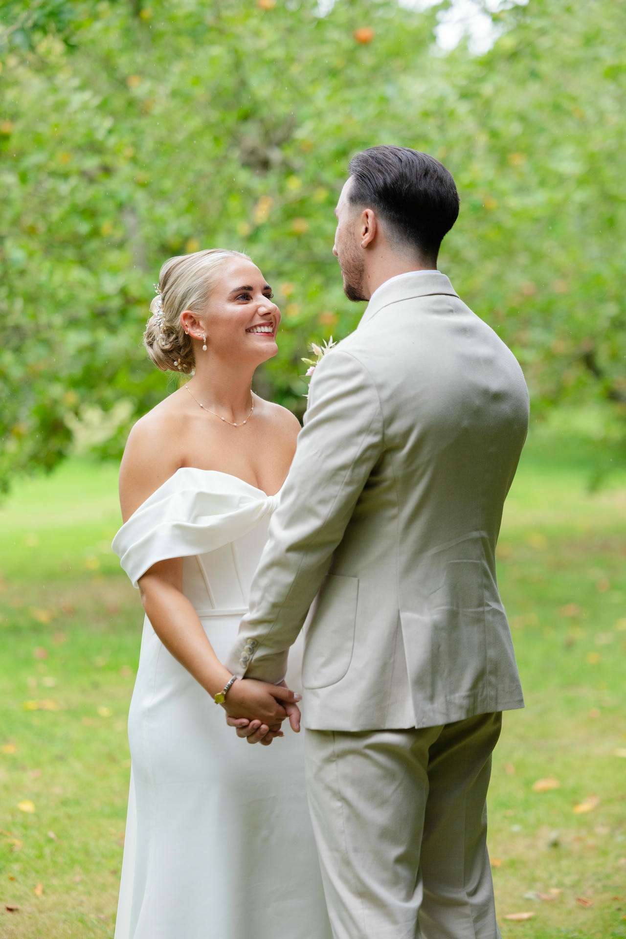 Bride and groom in wedding attire hold hands and smile at each other in a green outdoor setting.