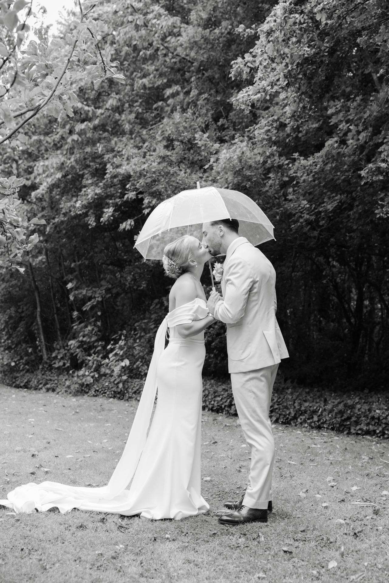 A bride in a long white wedding gown and a groom in a light suit kiss beneath a clear umbrella in a park.
