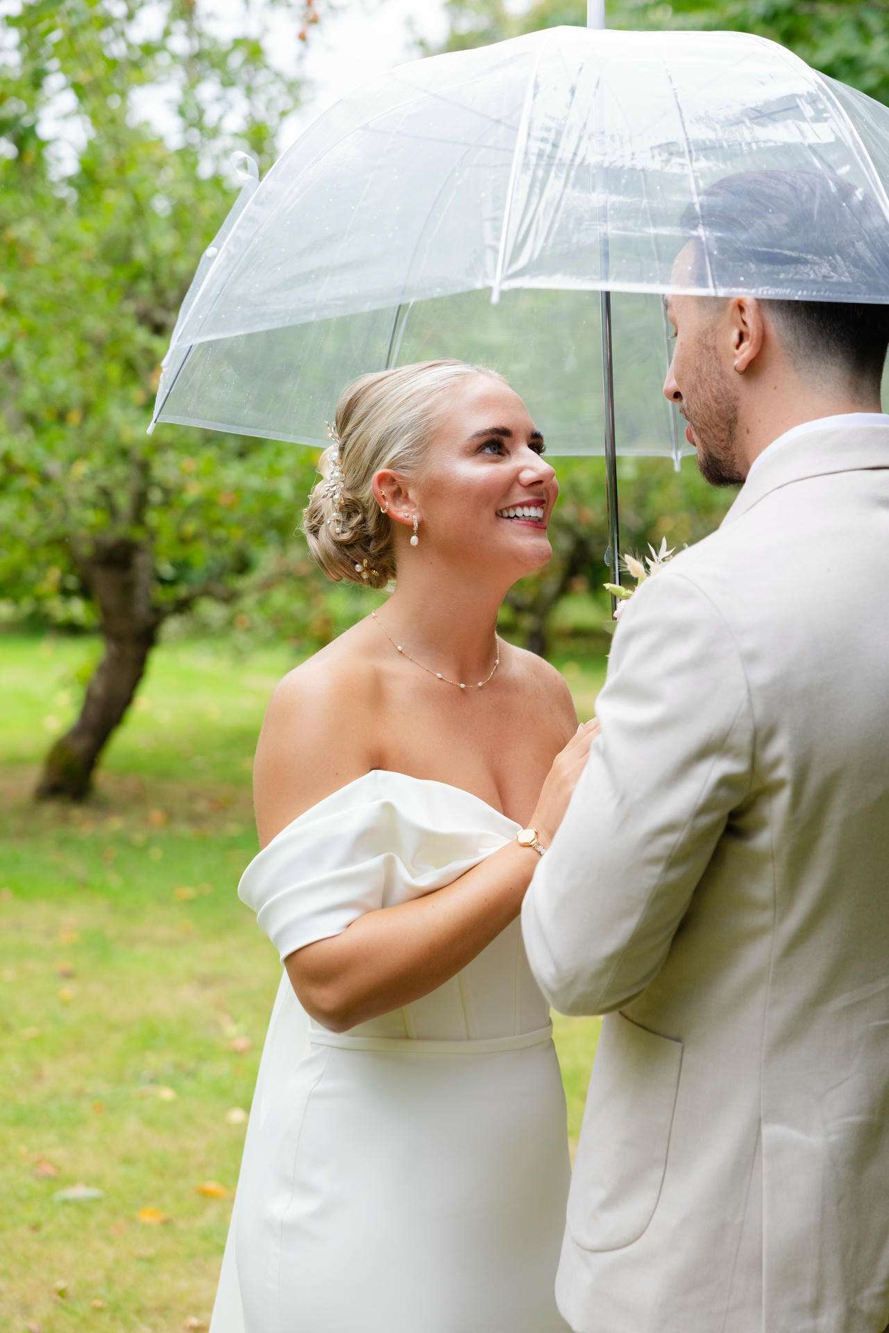 Bride in an off-the-shoulder white dress and groom in a light suit stand close under a clear umbrella in a lush outdoor garden.