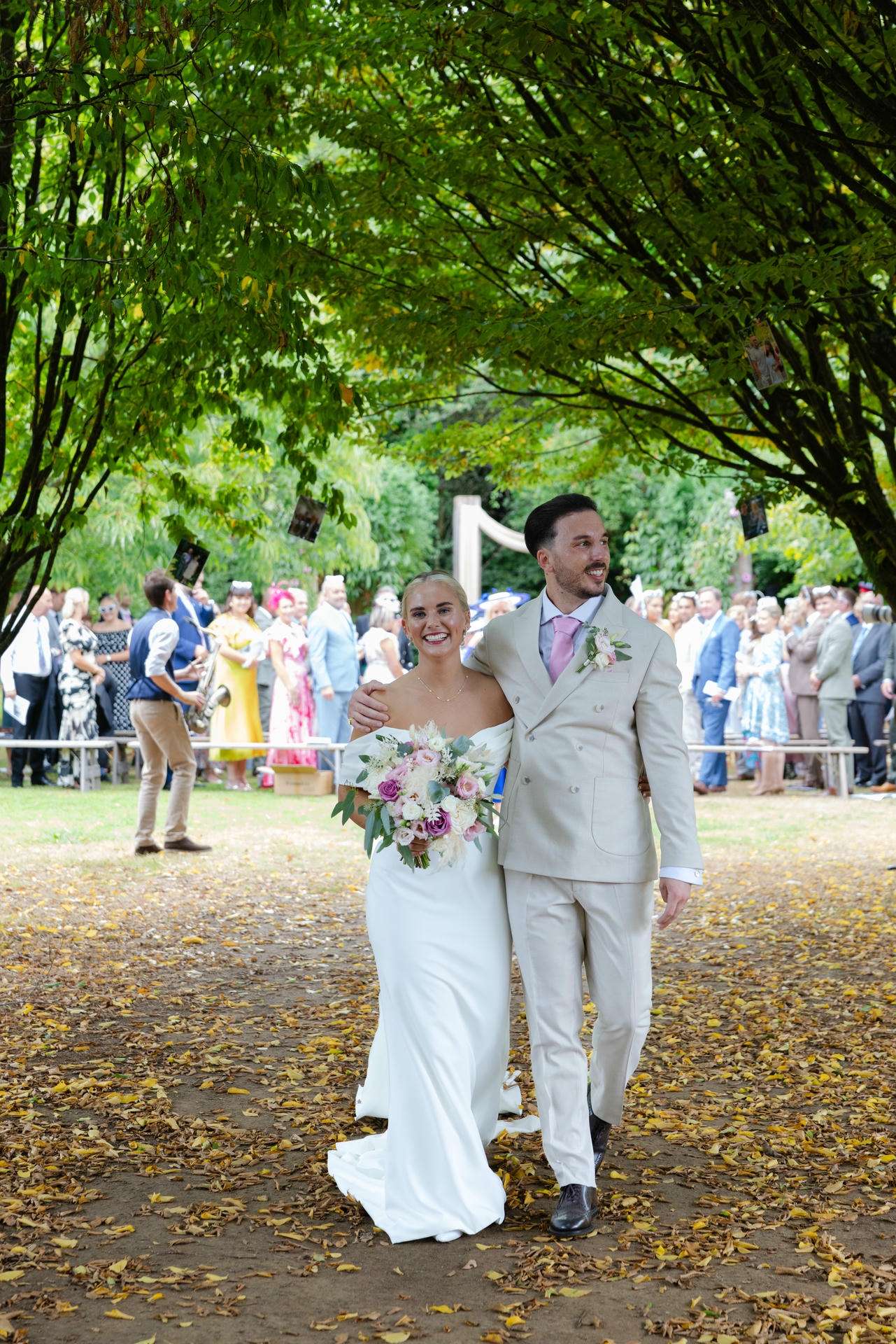Bride in a white gown and groom in a light suit walk arm in arm along a tree-lined outdoor wedding path, the bride holding a pink and white bouquet as guests look on.