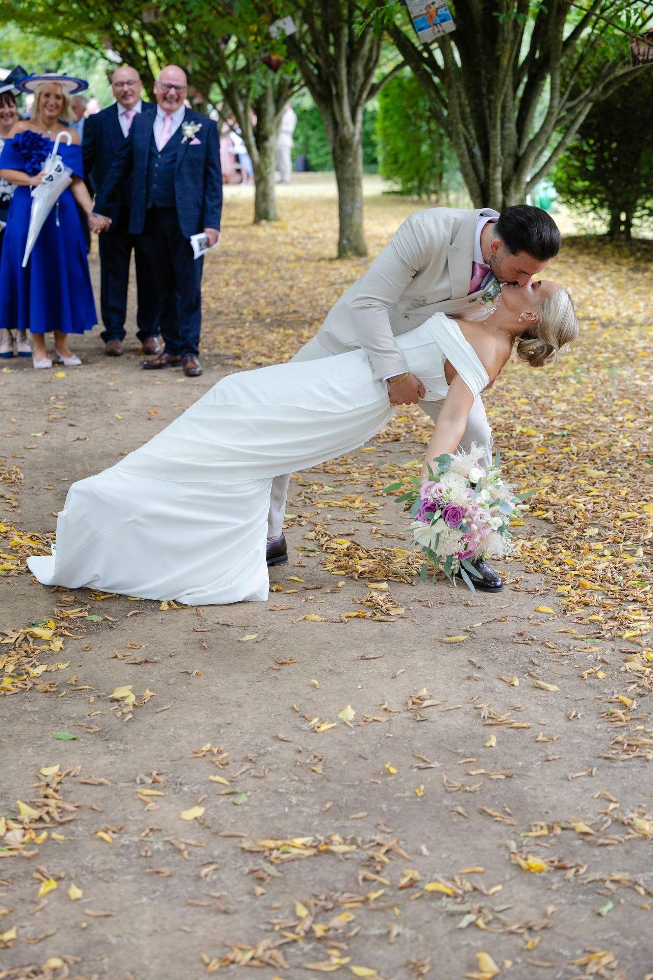 Groom in a light suit dips and kisses his bride in a white wedding dress as guests watch in a tree-lined park.