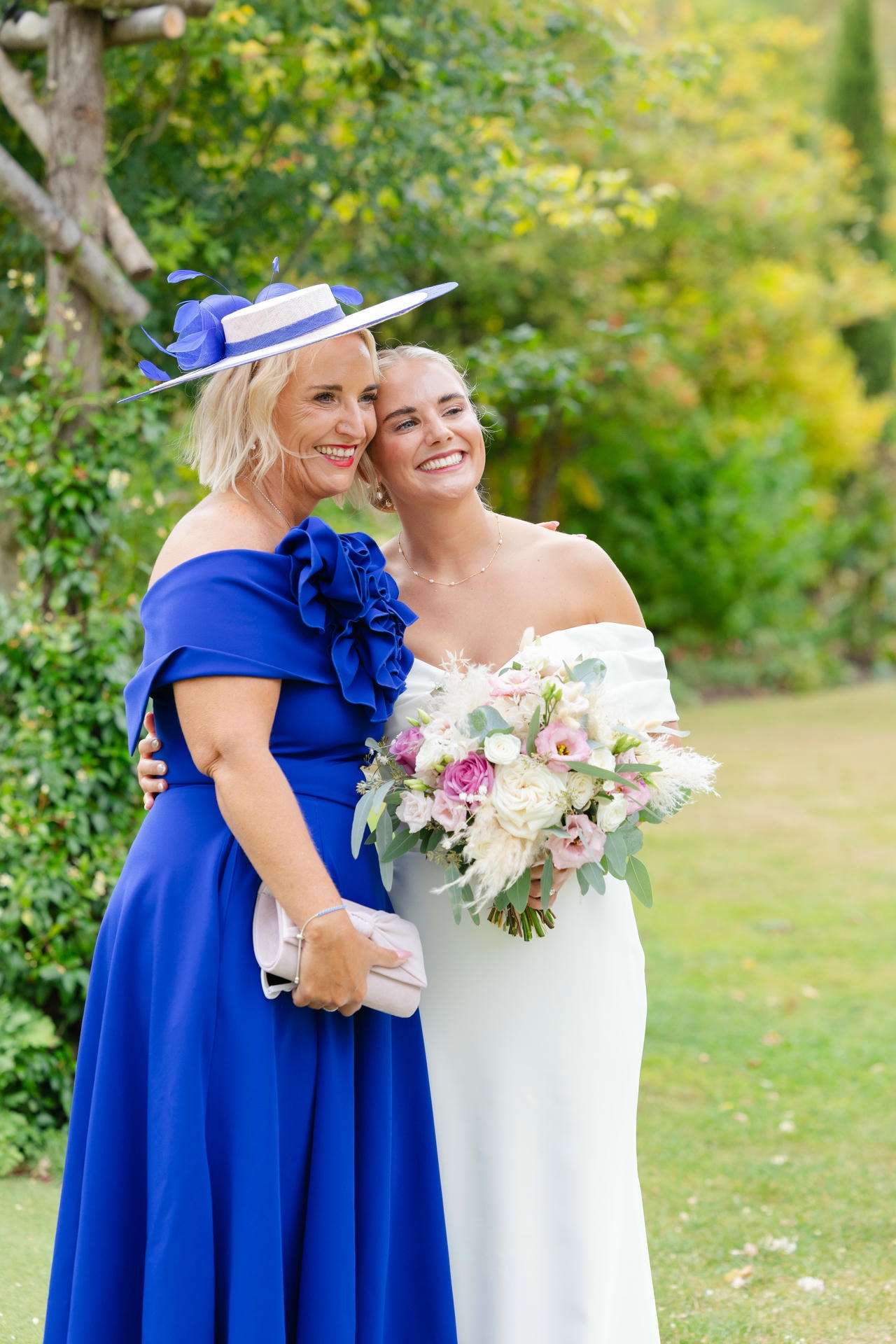 Smiling bride in a white off-shoulder gown holds a pastel bouquet while posing with a woman in a royal blue dress and matching hat in a lush garden.