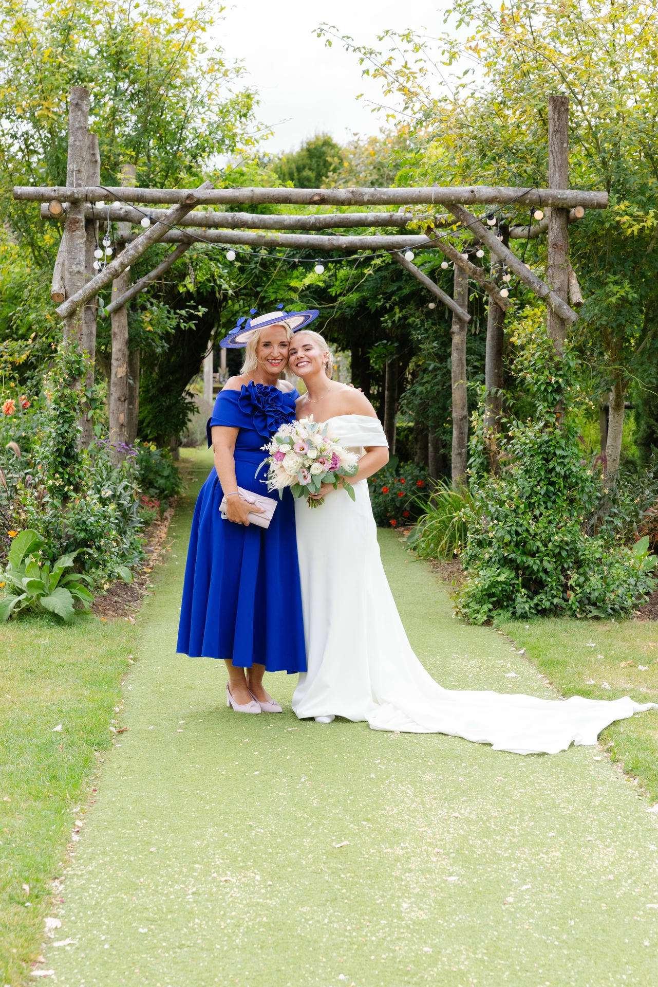 Bride in a white wedding dress holding a bouquet stands with a guest in a blue dress under a rustic wooden garden arch.