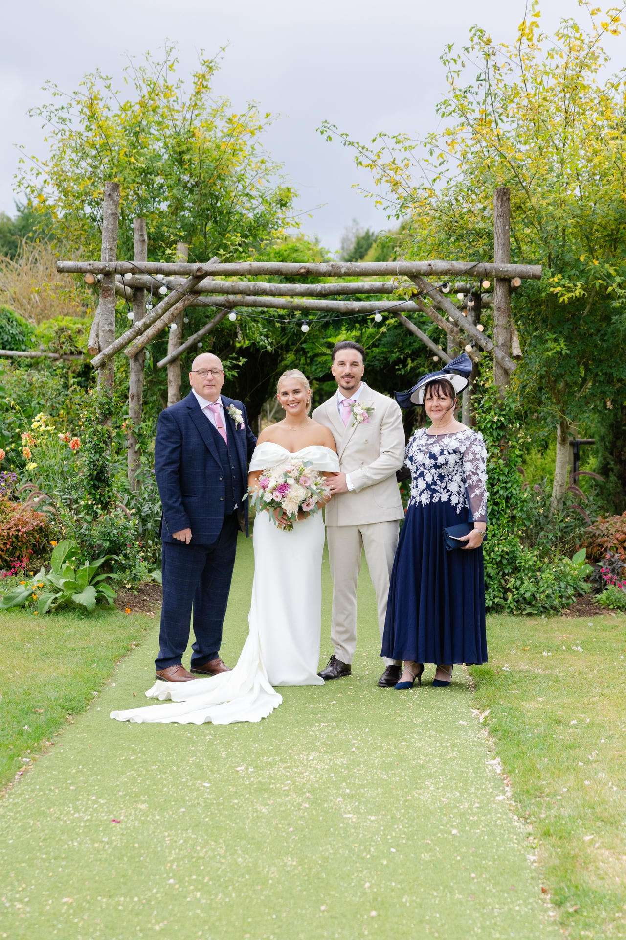 Bride and groom pose with two guests under a rustic wooden arch in a garden.