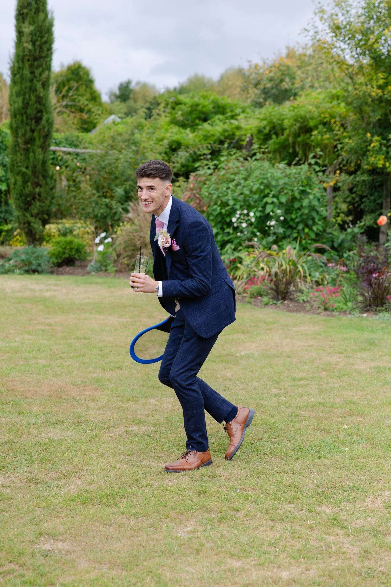 A man in a navy suit with a pink boutonniere smiles and leans forward while holding a drink in a garden with a blue hose looping behind him.