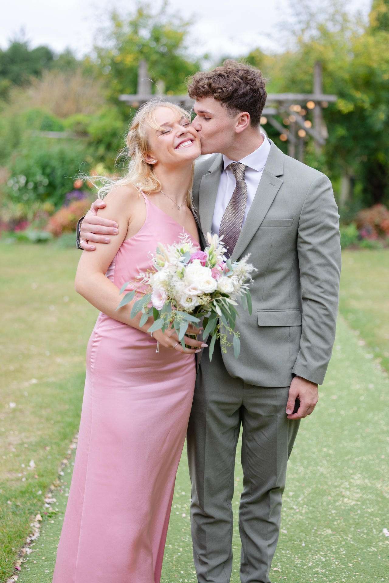 A man in a gray suit kisses a smiling woman in a pink dress who holds a bouquet of white and pink flowers in a garden.
