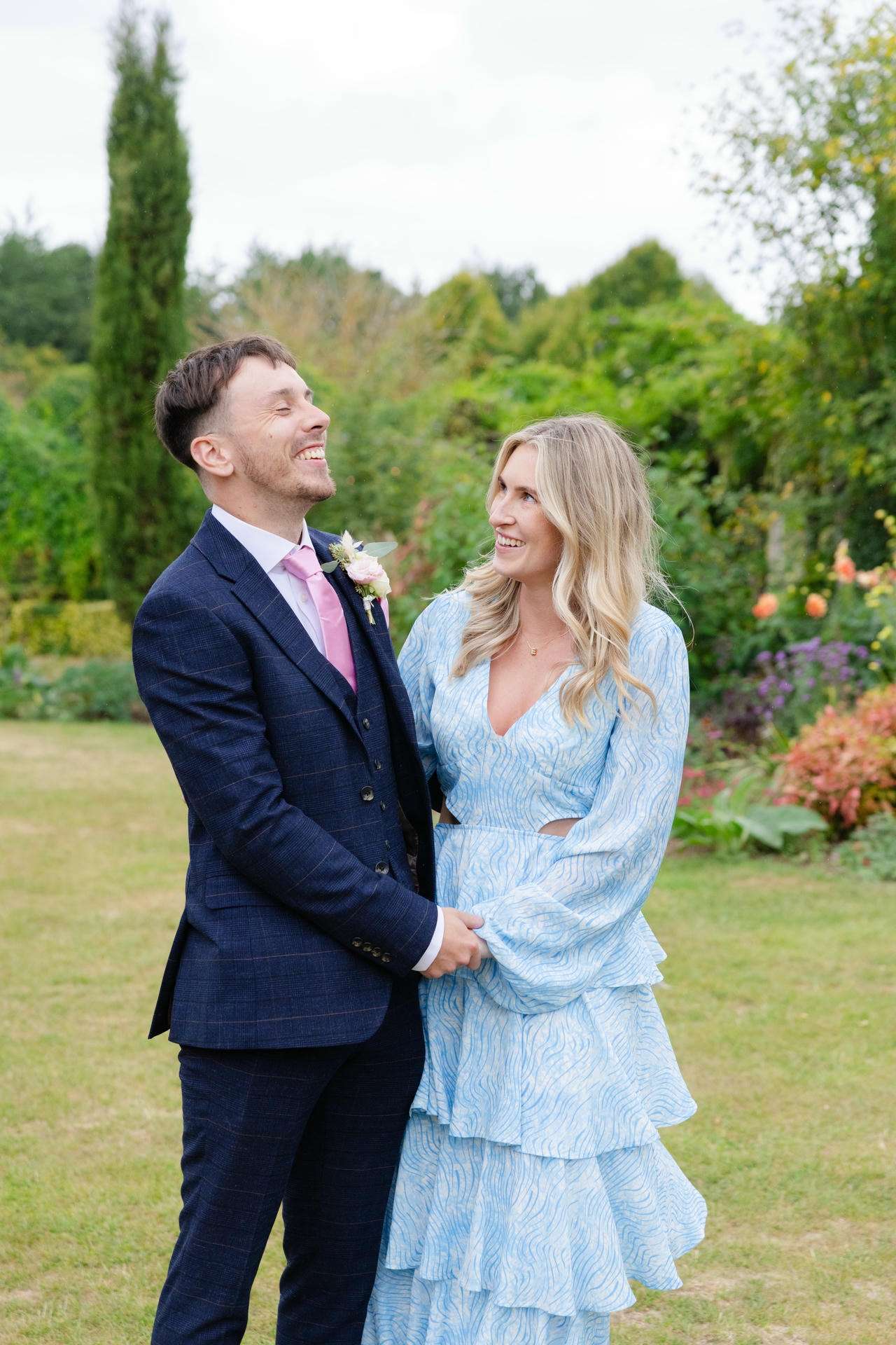 A couple stands in a garden, the man wearing a navy suit with a pink tie and boutonniere and the woman in a light blue, tiered dress, smiling at each other while holding hands.