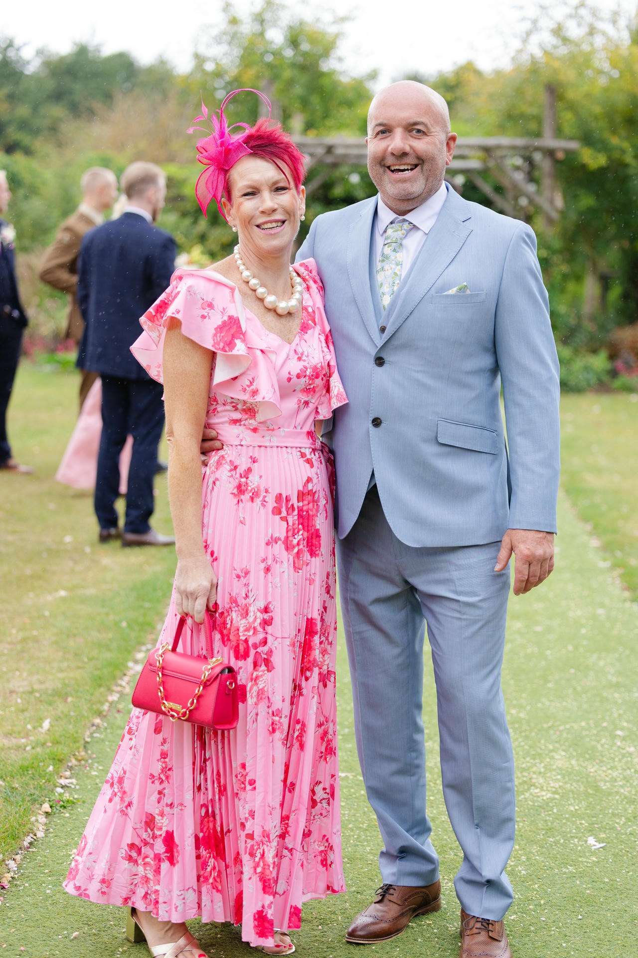 A smiling couple posing outdoors at a garden event; the woman wears a pink floral dress with a pink hat and pearl necklace, and the man wears a light blue suit.