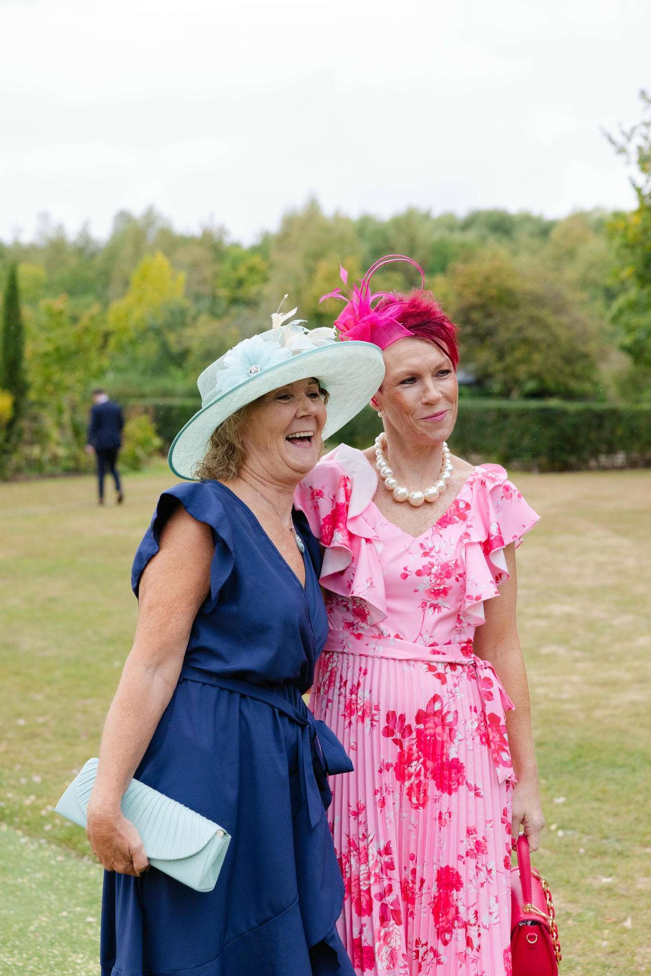 Two women in elegant dresses and hats pose on a grassy lawn, one wearing a navy dress with a light blue hat and the other in a pink floral dress with a pink fascinator.