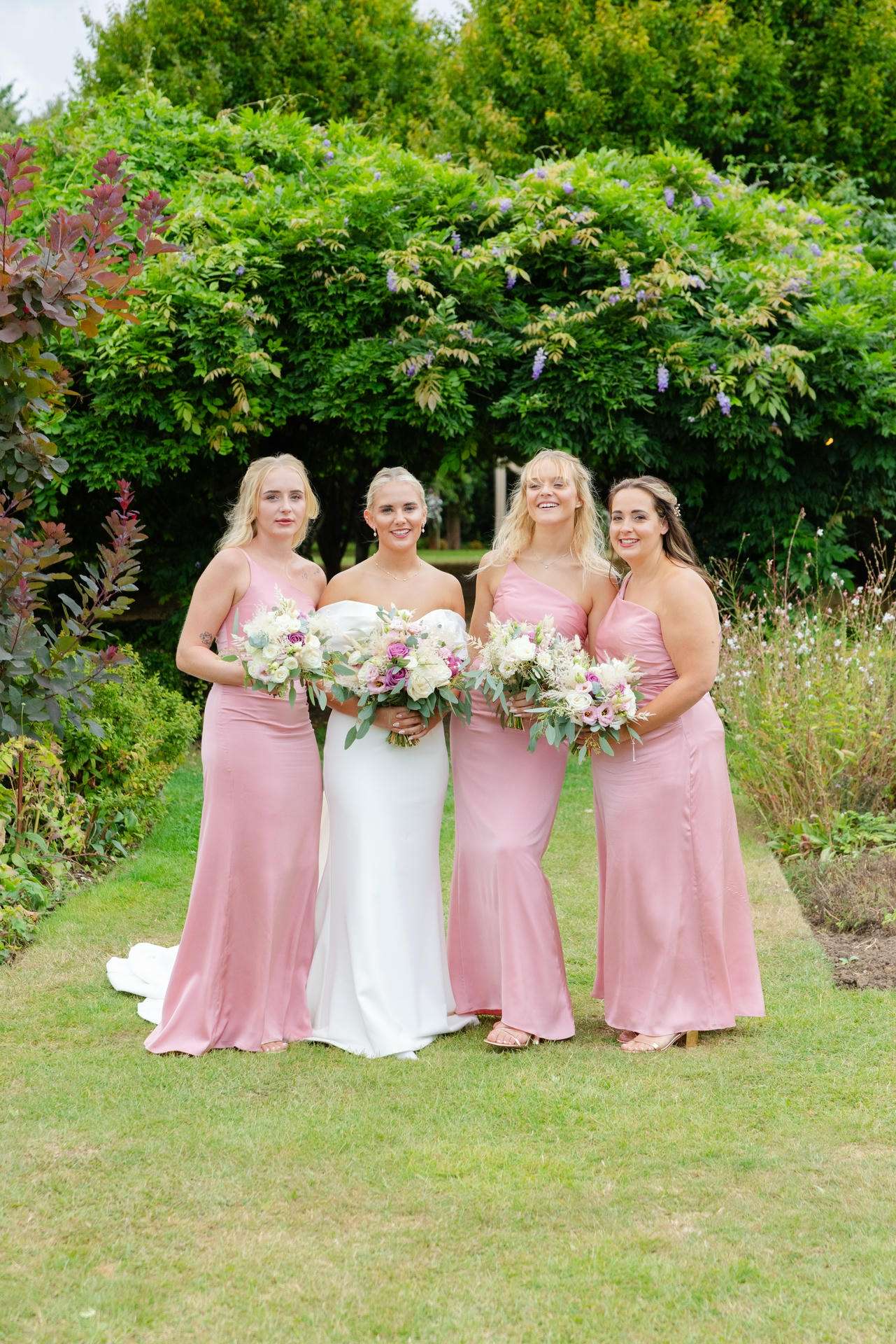 Bride in a white wedding gown stands with three bridesmaids in pink dresses, each holding bouquets, in a lush garden.