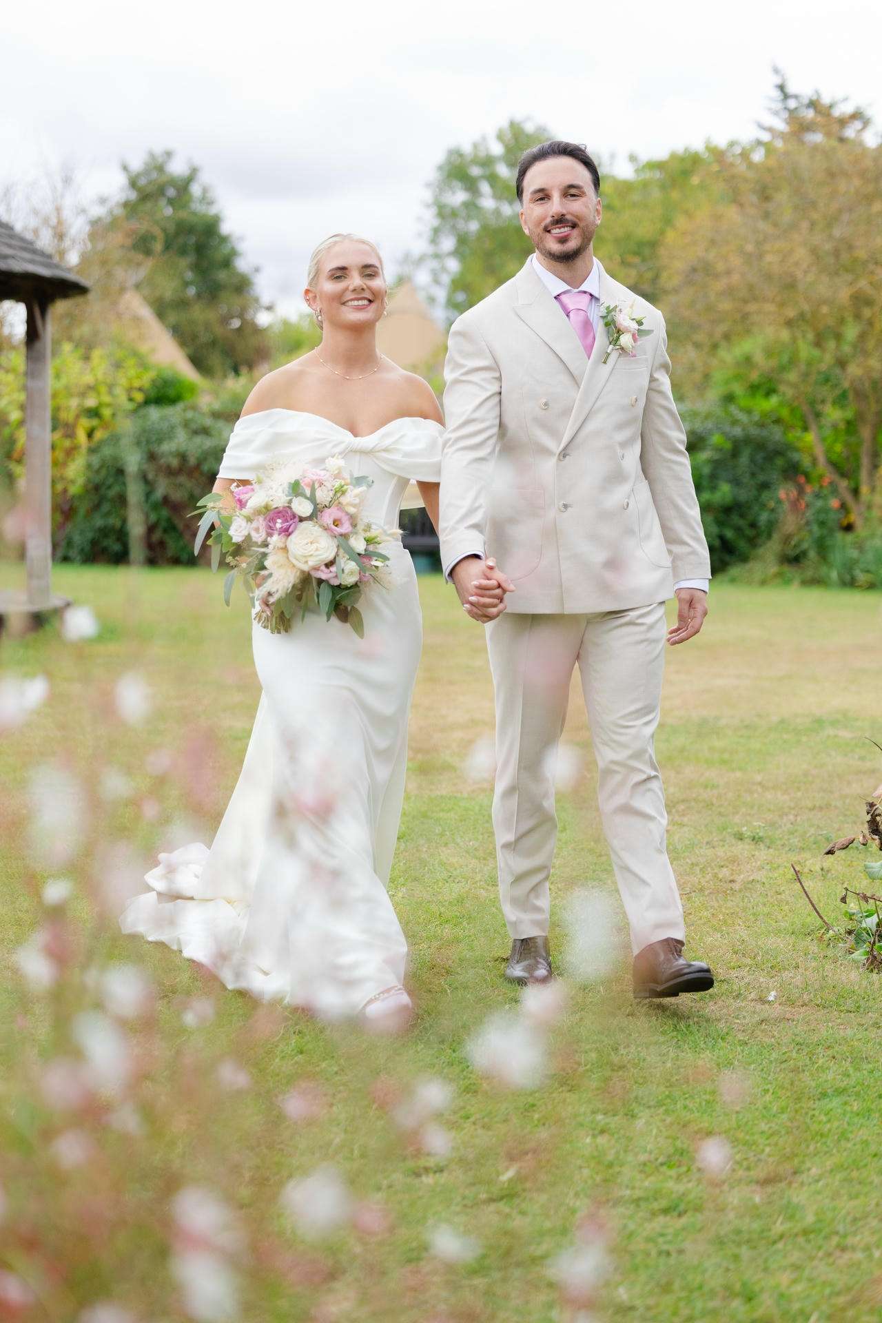 A bride in an off-the-shoulder white wedding dress and a groom in a light beige suit walk hand in hand through a garden, smiling, with the bride holding a pink and white bouquet at The Gardens Wedding venue