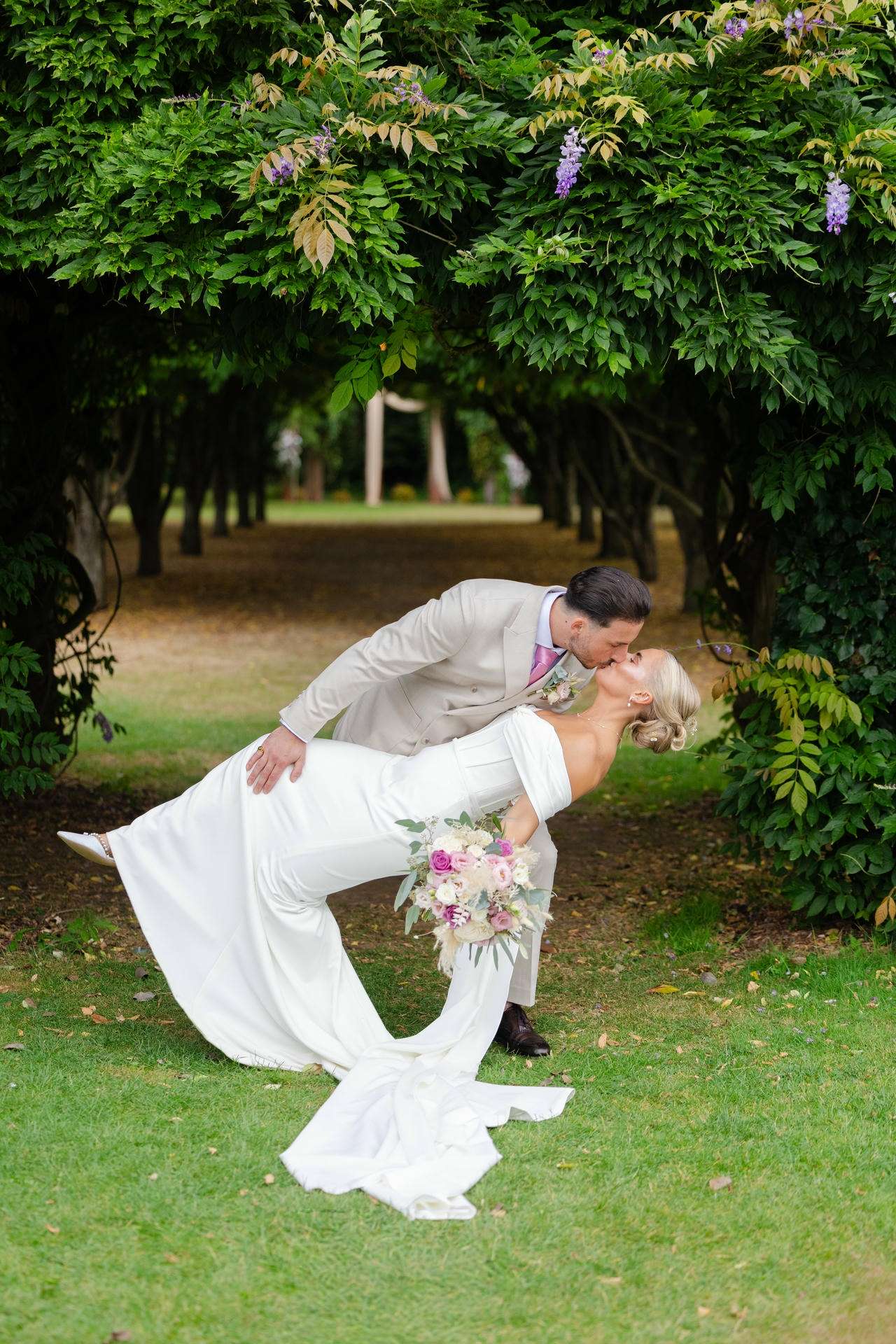 Bride and groom kiss as he dips her under a lush green arch, the bride holding a pink and white bouquet at The Gardens Wedding venue