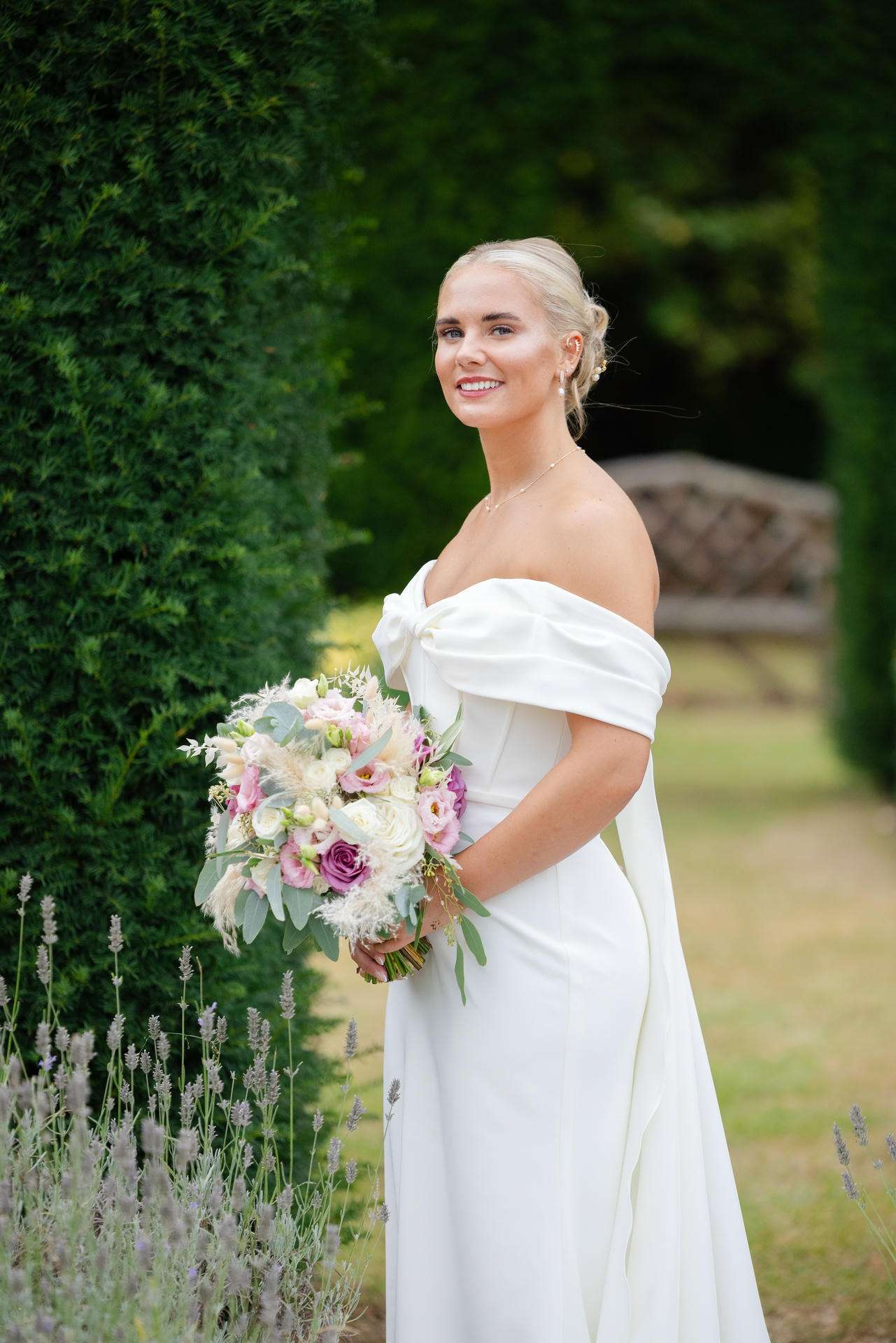 A smiling blonde bride in an off-the-shoulder white wedding dress holds a pastel bouquet outdoors at The Gardens Wedding venue